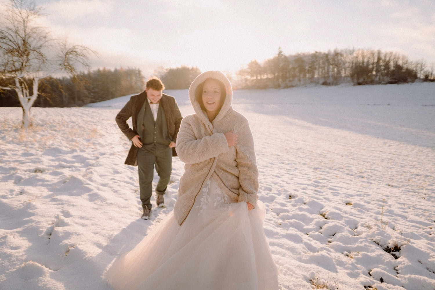 Ein Paar geht durch ein verschneites Feld in der Nähe von Hammerschloss Stodl Schmidmühlen. Die Frau trägt ein weißes Brautkleid und einen beigen Mantel, der Mann einen dunklen Anzug. Sie sind umgeben von einer ruhigen, sonnenbeschienenen Winterlandschaft mit kahlen Bäumen und einem sanften Schimmer am Himmel.