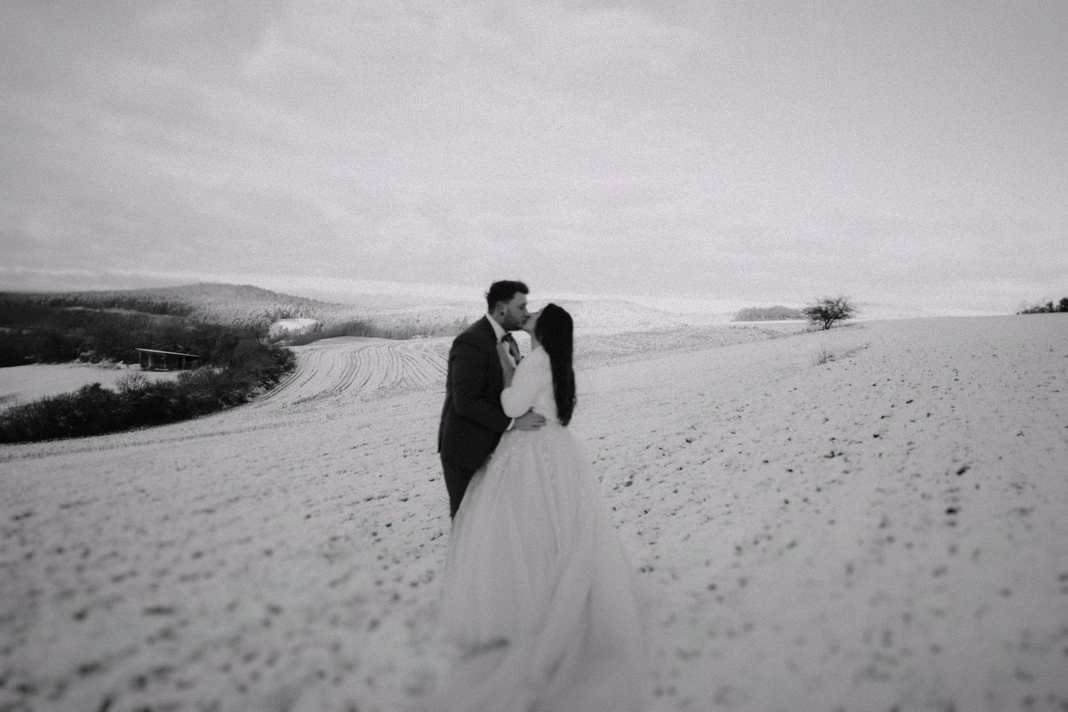 Umarmend auf einem schneebedeckten Feld in der Nähe von Hammerschloss Stodl blickt sich ein Paar an. Die weite Landschaft zeigt sanfte Hügel und einen einsamen Baum unter einem bewölkten Himmel. Die in Schwarzweiß festgehaltene Szene ruft eine heitere und romantische Atmosphäre hervor, die an den zeitlosen Charme von Schmidmühlen erinnert.