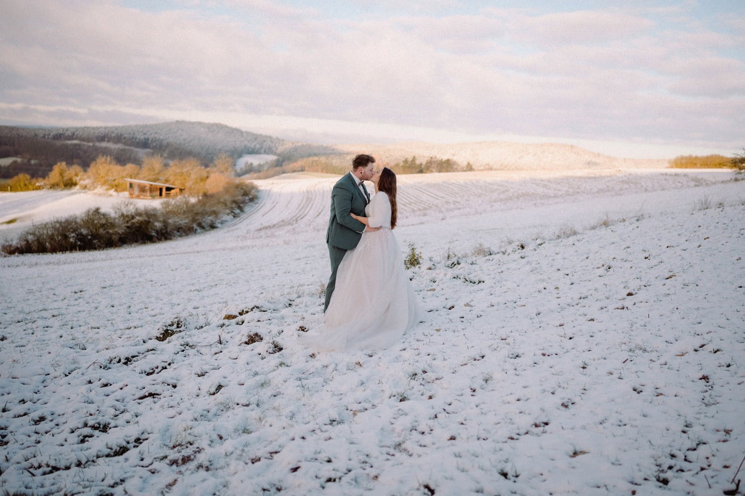 Ein Paar umarmt sich auf einem schneebedeckten Feld unter einem teilweise bewölkten Himmel. Die Landschaft besteht aus sanften Hügeln, einem kleinen Holzbau, der an den rustikalen Charme von Hammerschloss Stodl erinnert, und in der Ferne schneebedeckten Bäumen. Sie scheinen einen romantischen Moment in der ruhigen Winterlandschaft zu genießen.