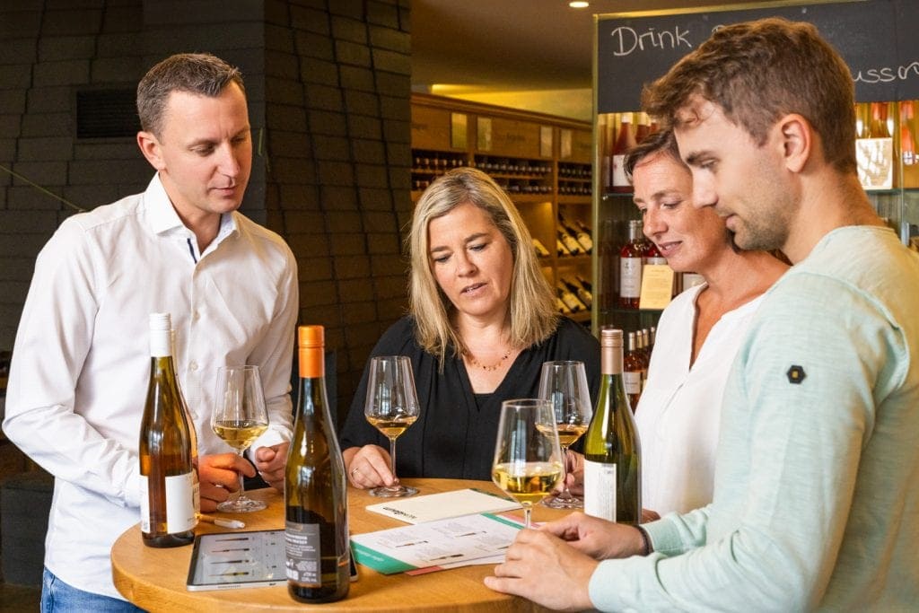 Gruppe von Menschen beim Weinverkostungsevent in einer Bar, Weinflaschen und Gläser im Vordergrund.