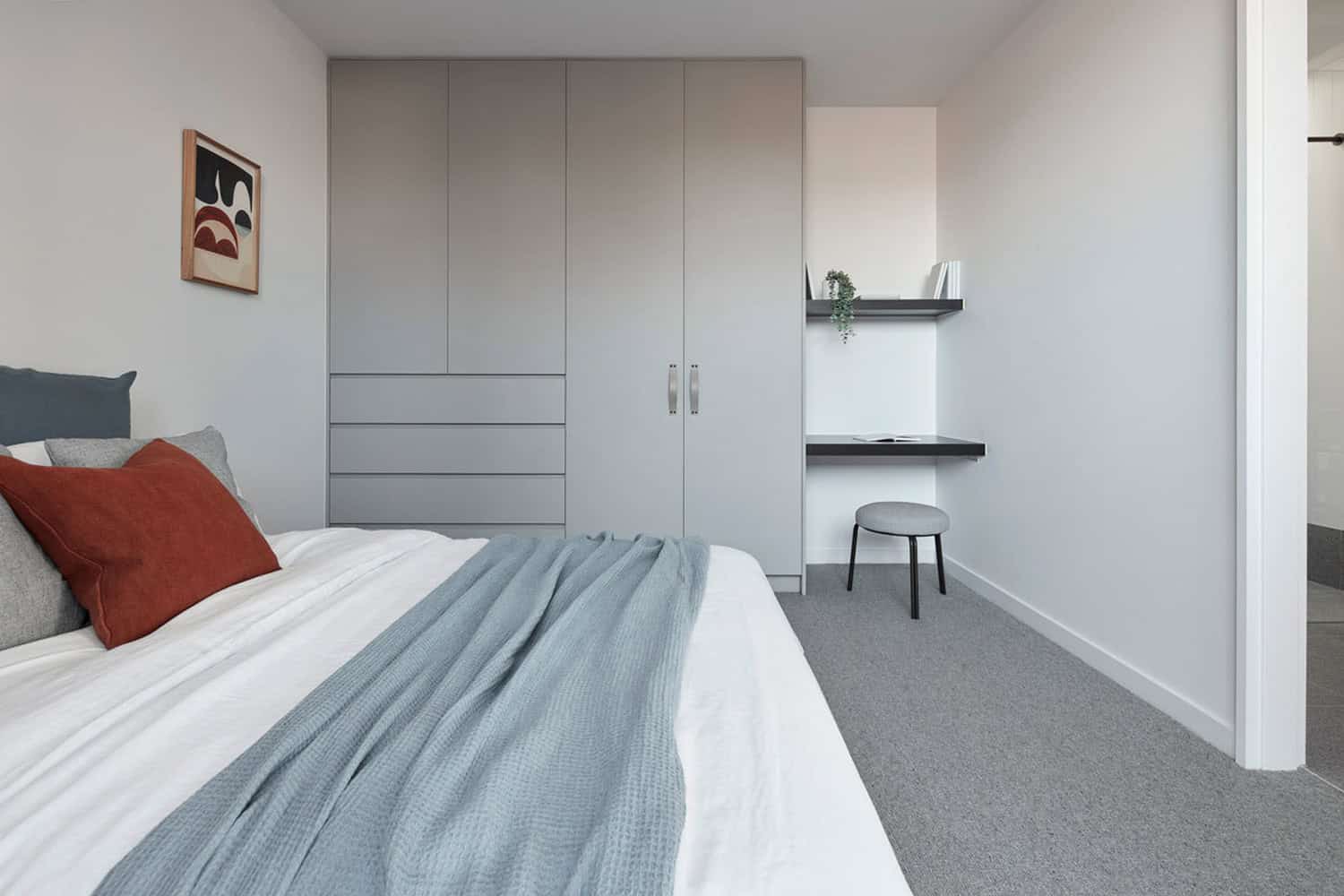 Modern bedroom featuring grey built-in wardrobes, a compact study nook with stool, and bed styled in blue and rust.