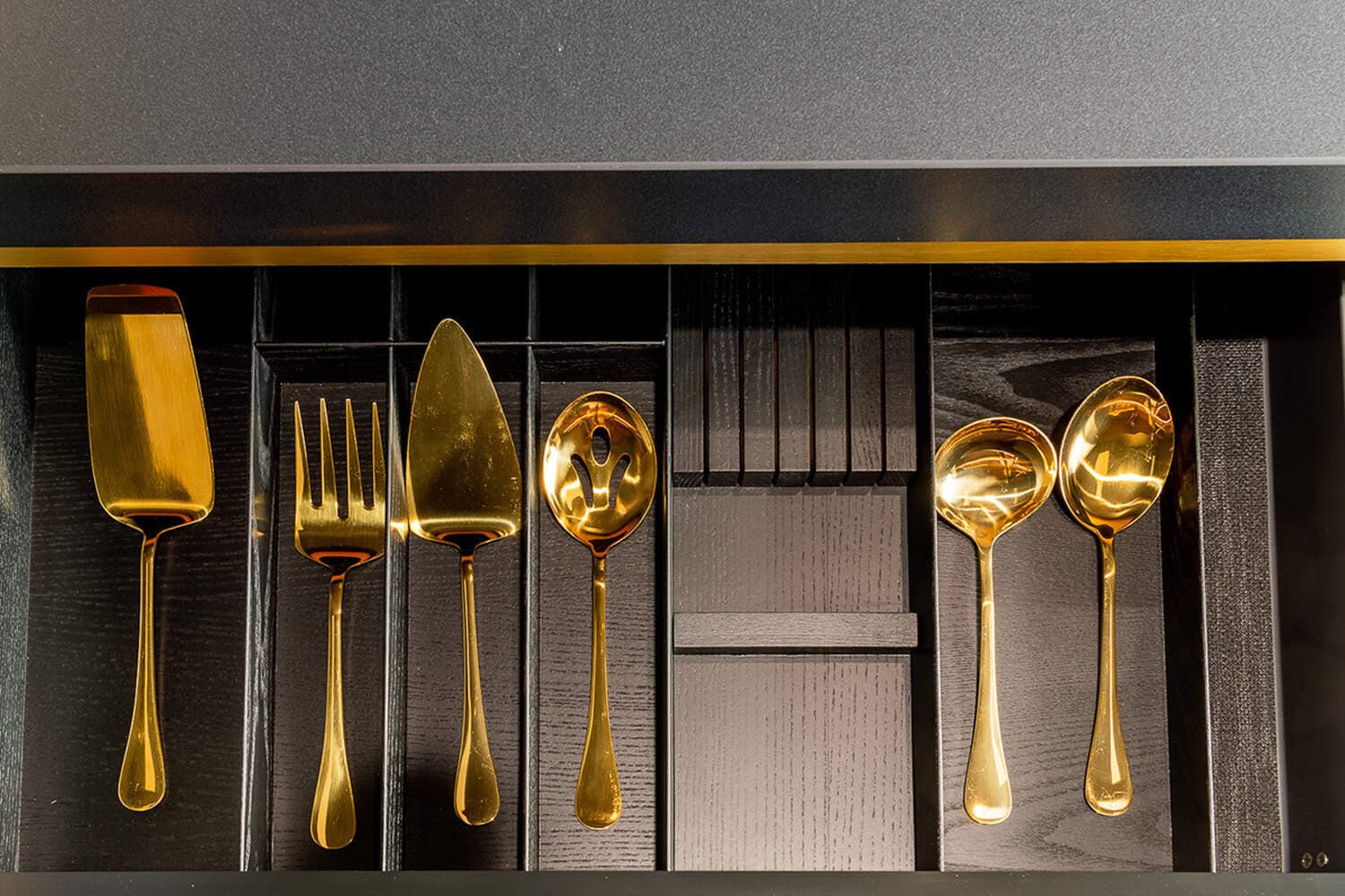 Gold serving utensils and cutlery arranged in a dark wood drawer tray divider.