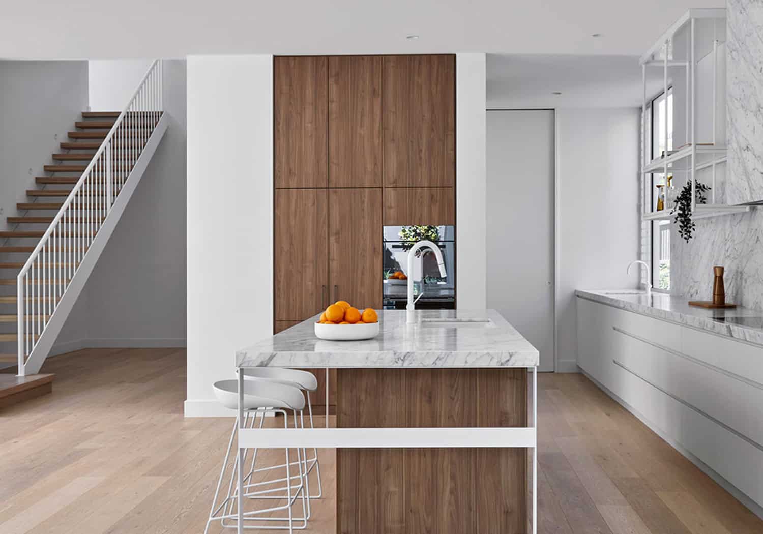 Open-plan kitchen with marble island, dark wood cabinetry, white-railed staircase, and timber flooring.