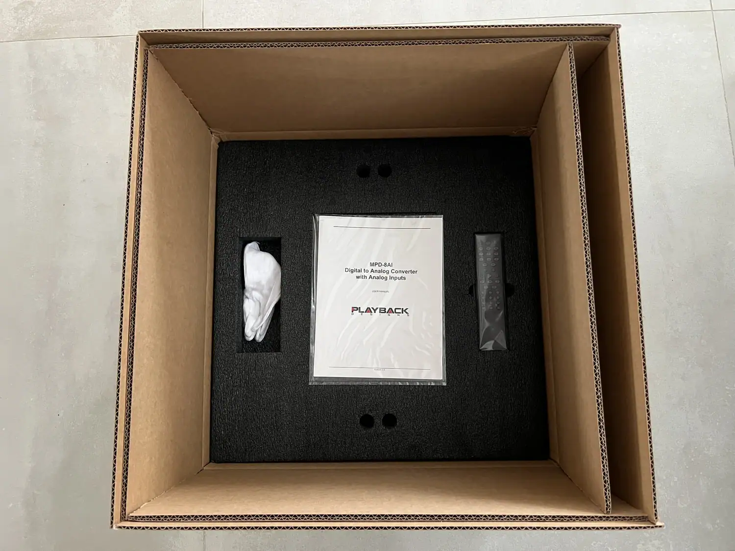 Carton du Playback Designs MPD-8AI ouvert avec des gants blancs, la documentation et la télécommande en aluminium