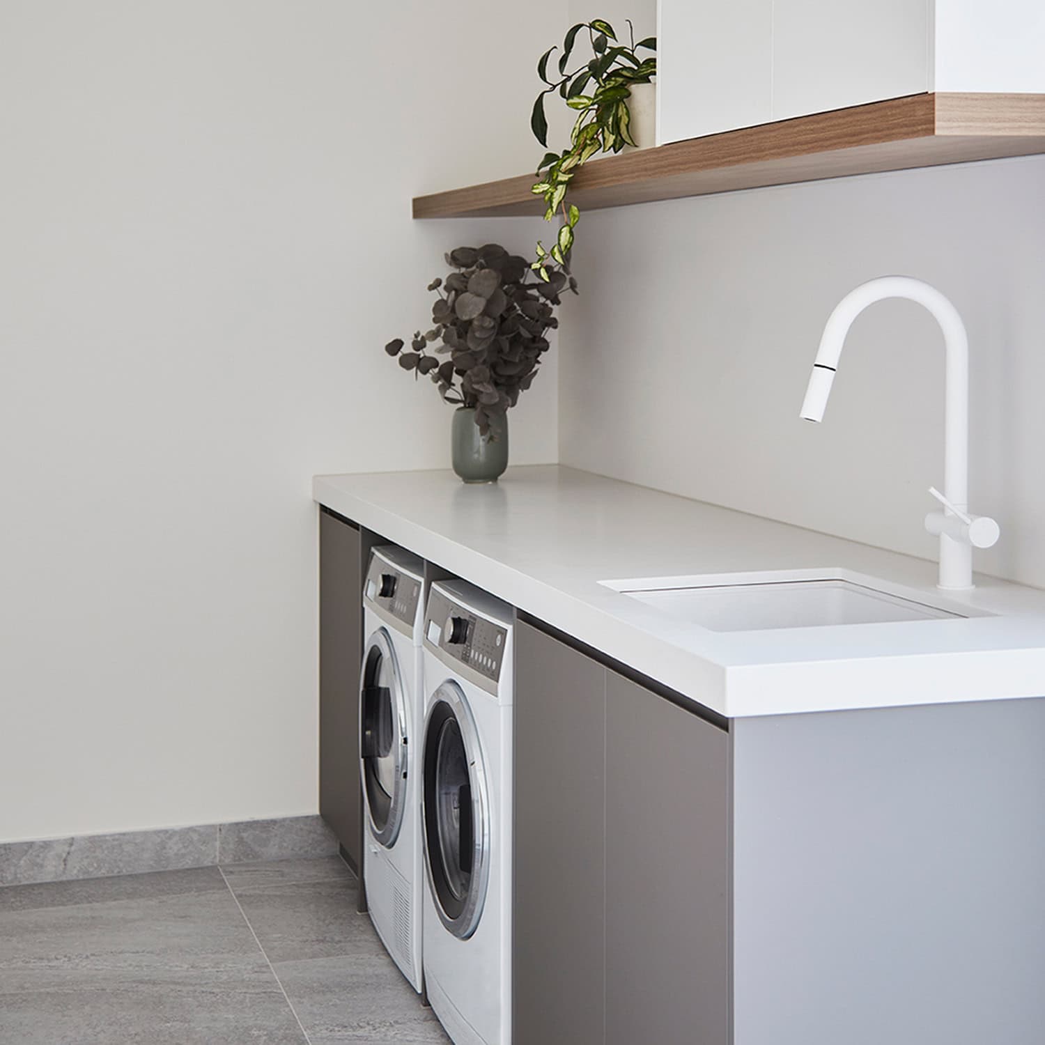 Sleek modern laundry room with white gooseneck tap, stone benchtop, grey cabinets, and washer-dryer set.