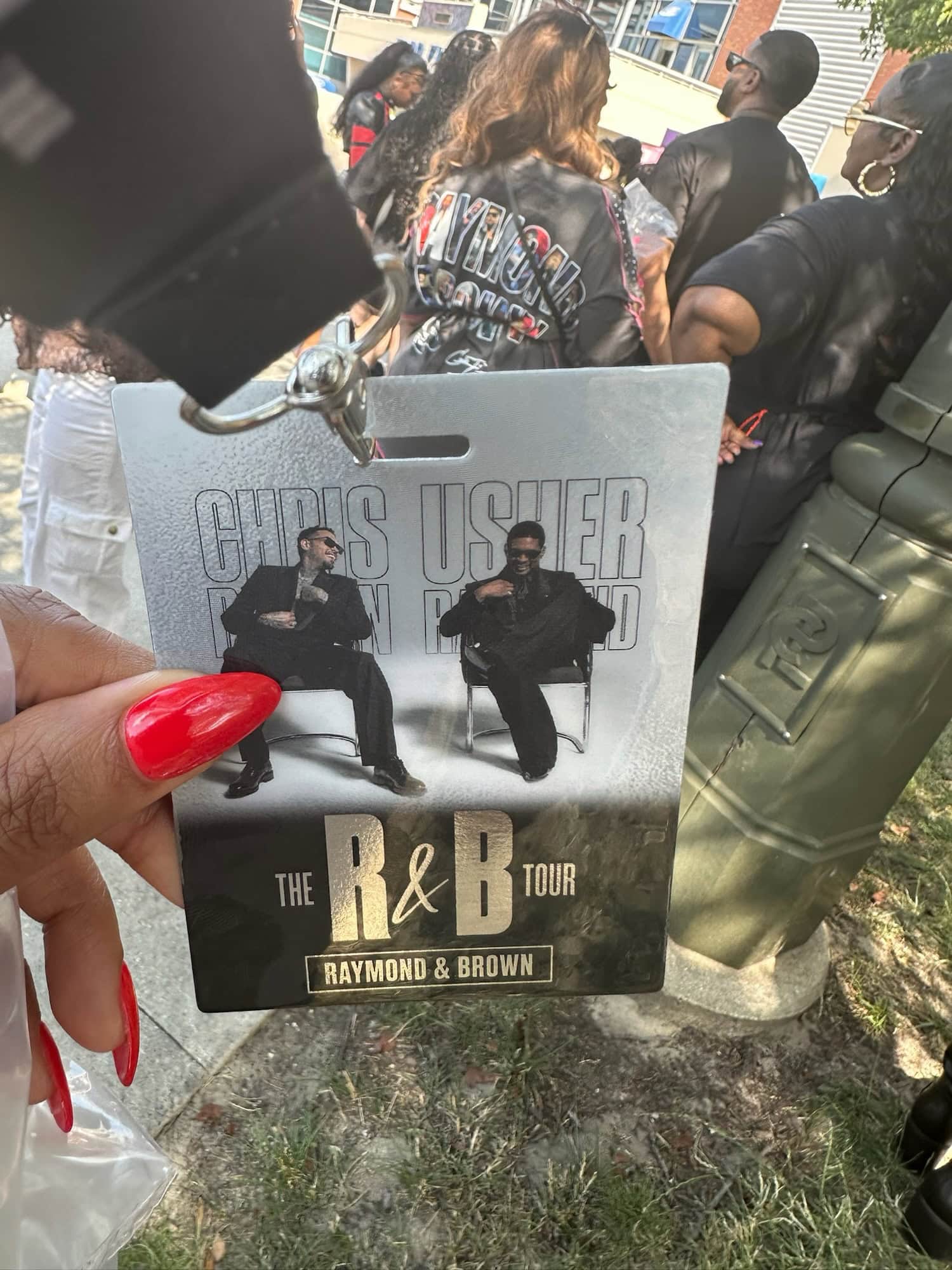 Hand with long red nails holding a Raymond & Brown R&B Tour VIP pass while waiting outside Ford Field in Detroit.