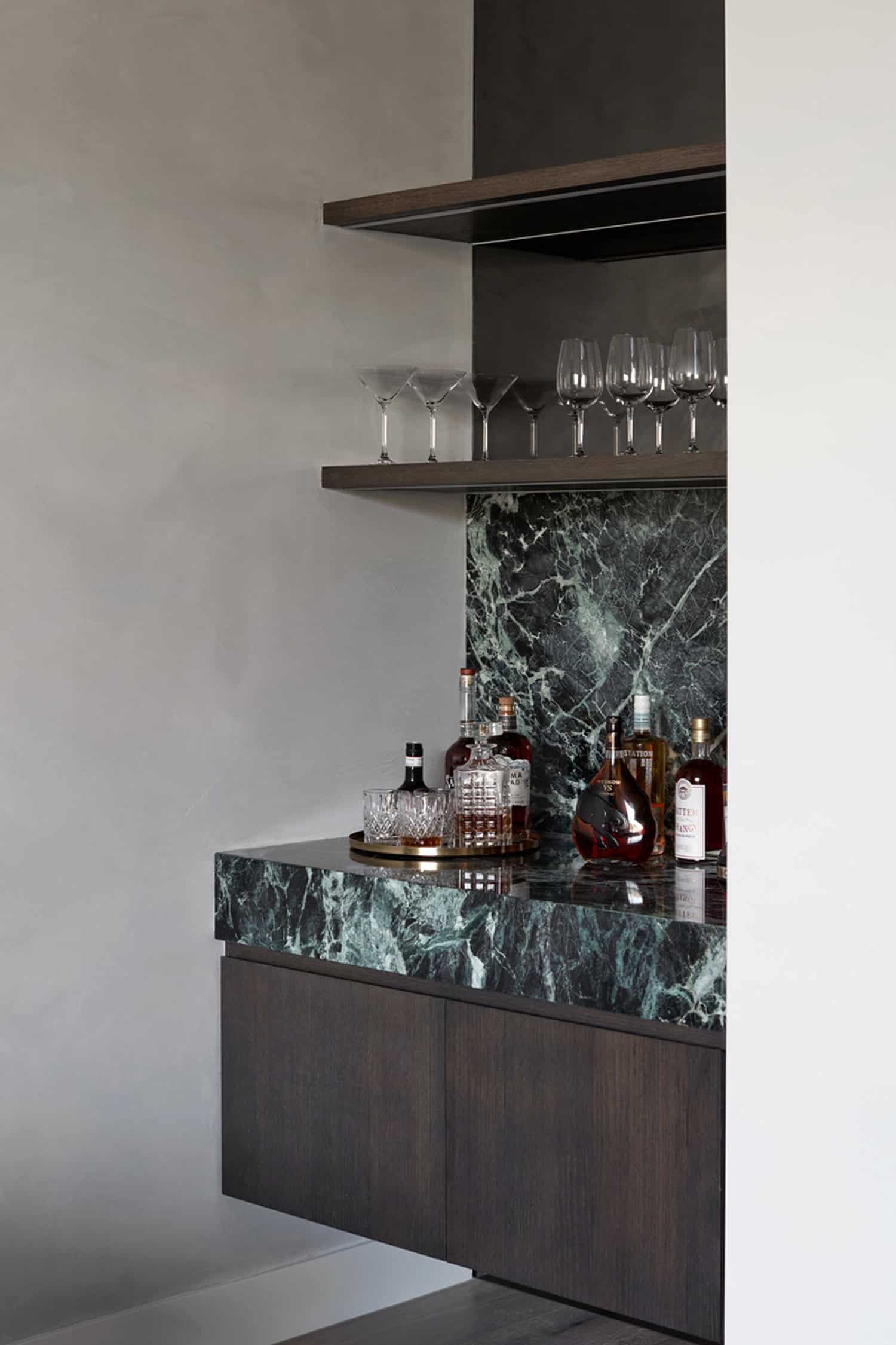 Built-in home bar with deep green marble countertop, dark wood cabinets, and floating glassware shelves.