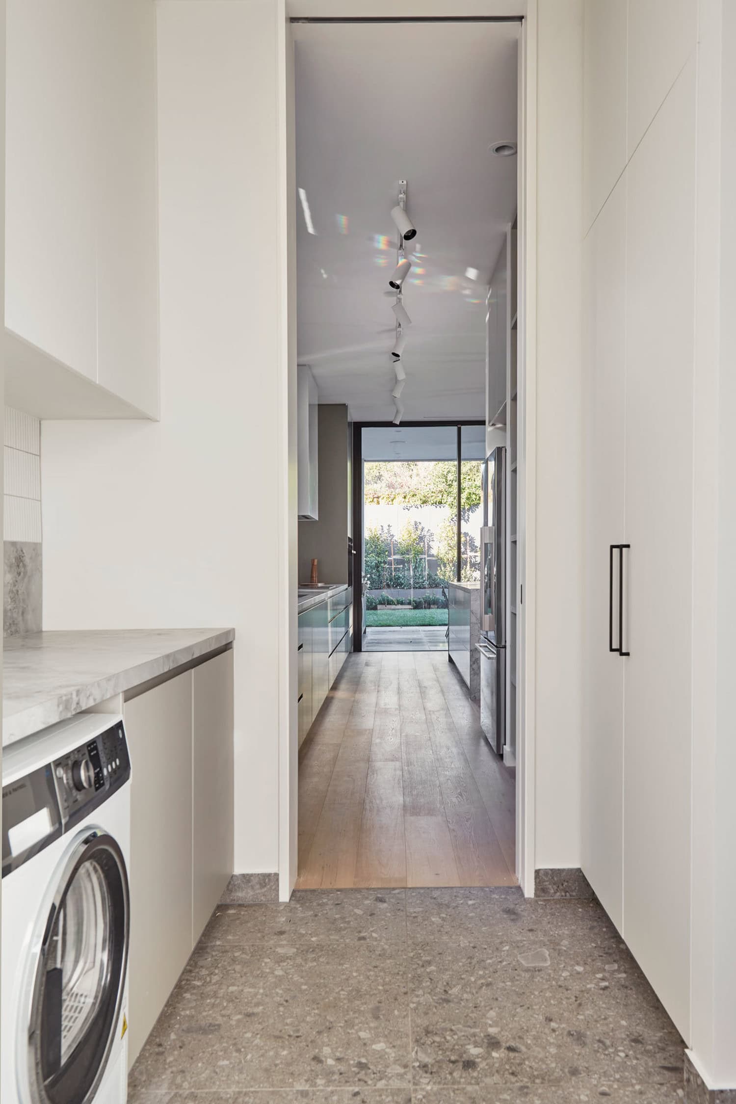 View from modern laundry room leading down hallway into light-filled open kitchen and garden beyond.