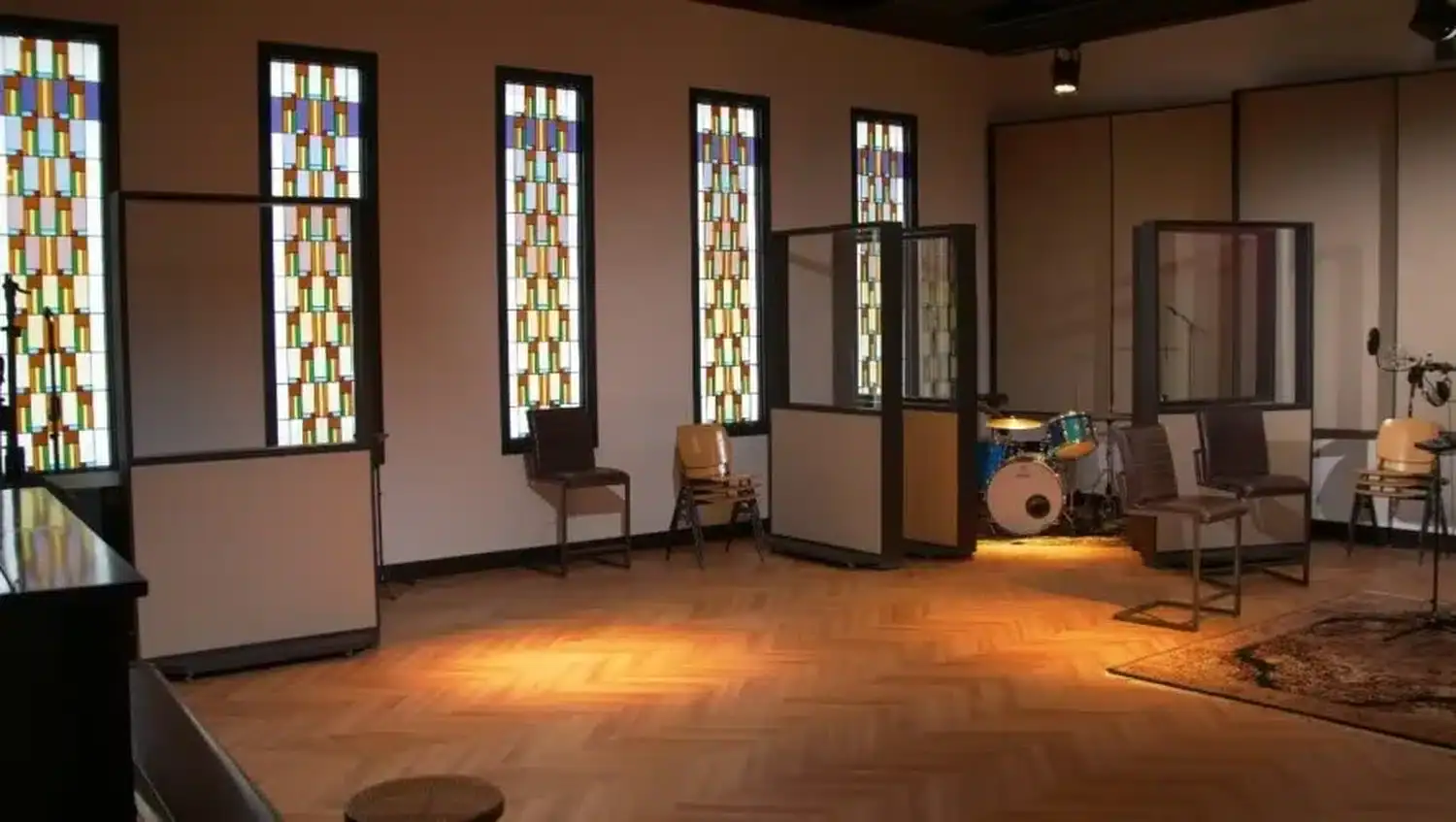Intérieur d'une pièce avec des vitraux colorés, du parquet, une batterie Alpha Audio et diverses chaises éparpillées (par 1877.audio)