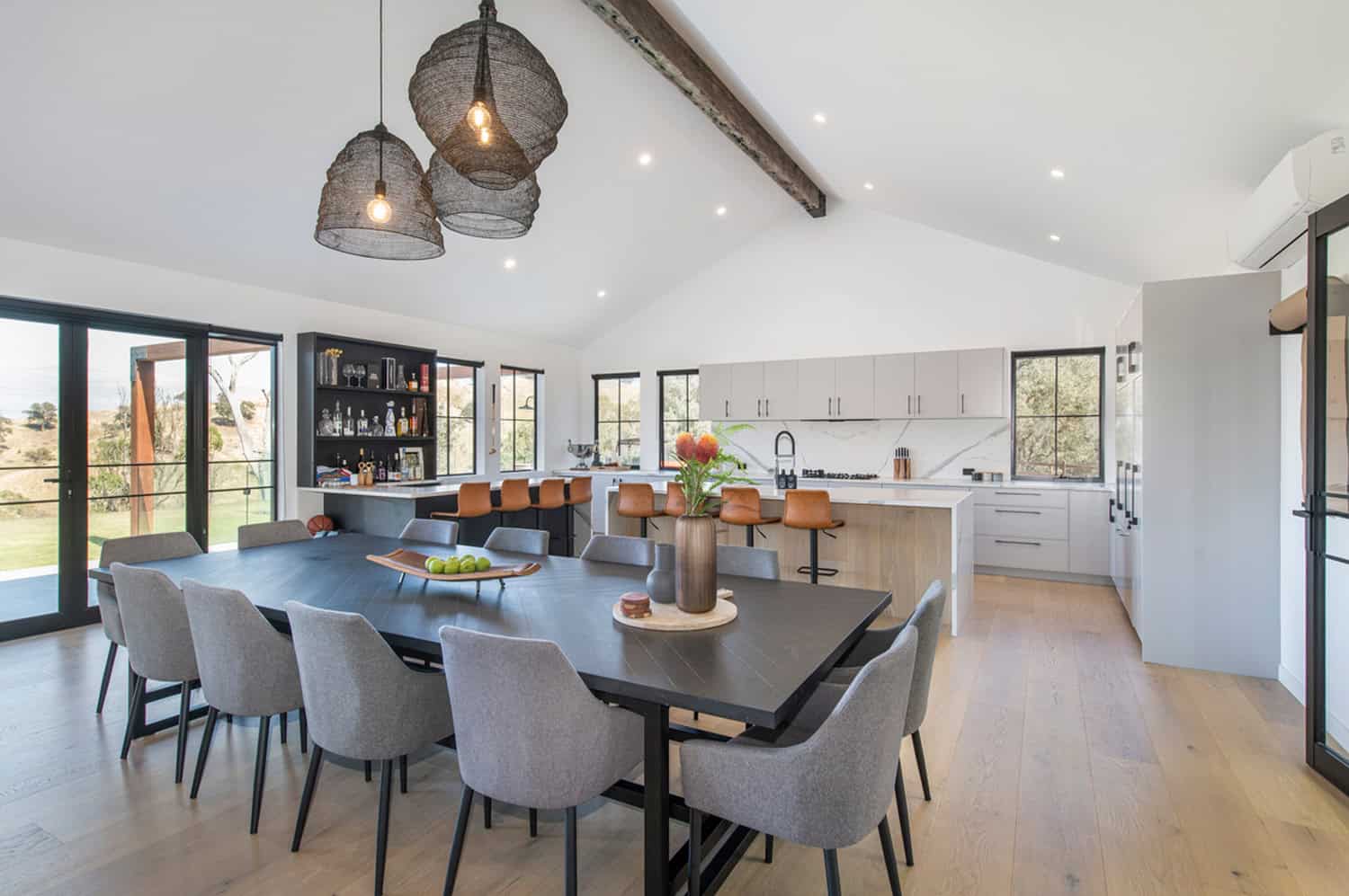 Open-plan kitchen and bar area with marble island, tan stools, dark liquor cabinet, and timber beam ceiling.
