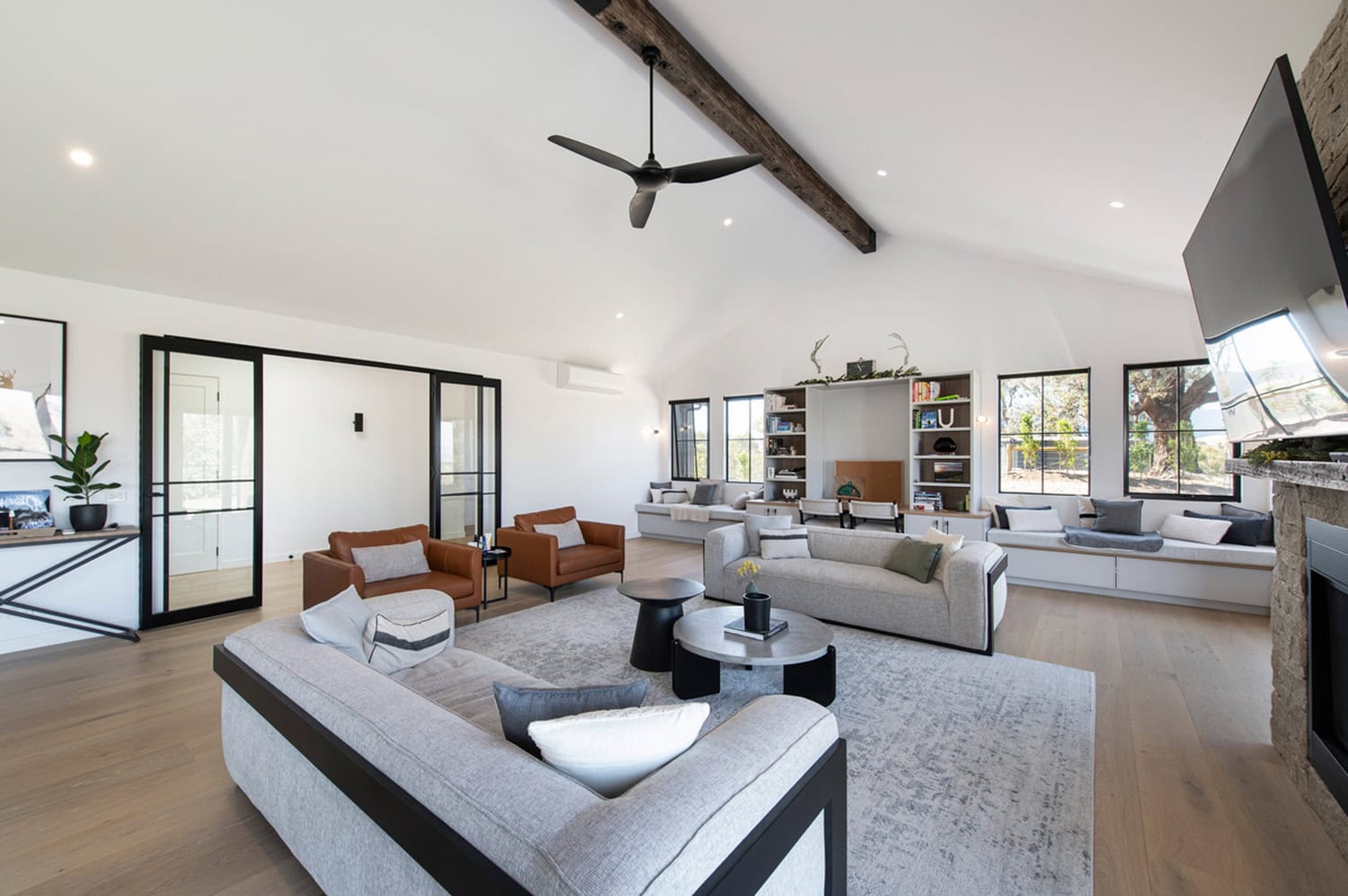 Spacious open living room with grey sofas, brown leather armchairs, exposed beam ceiling, and window seats.