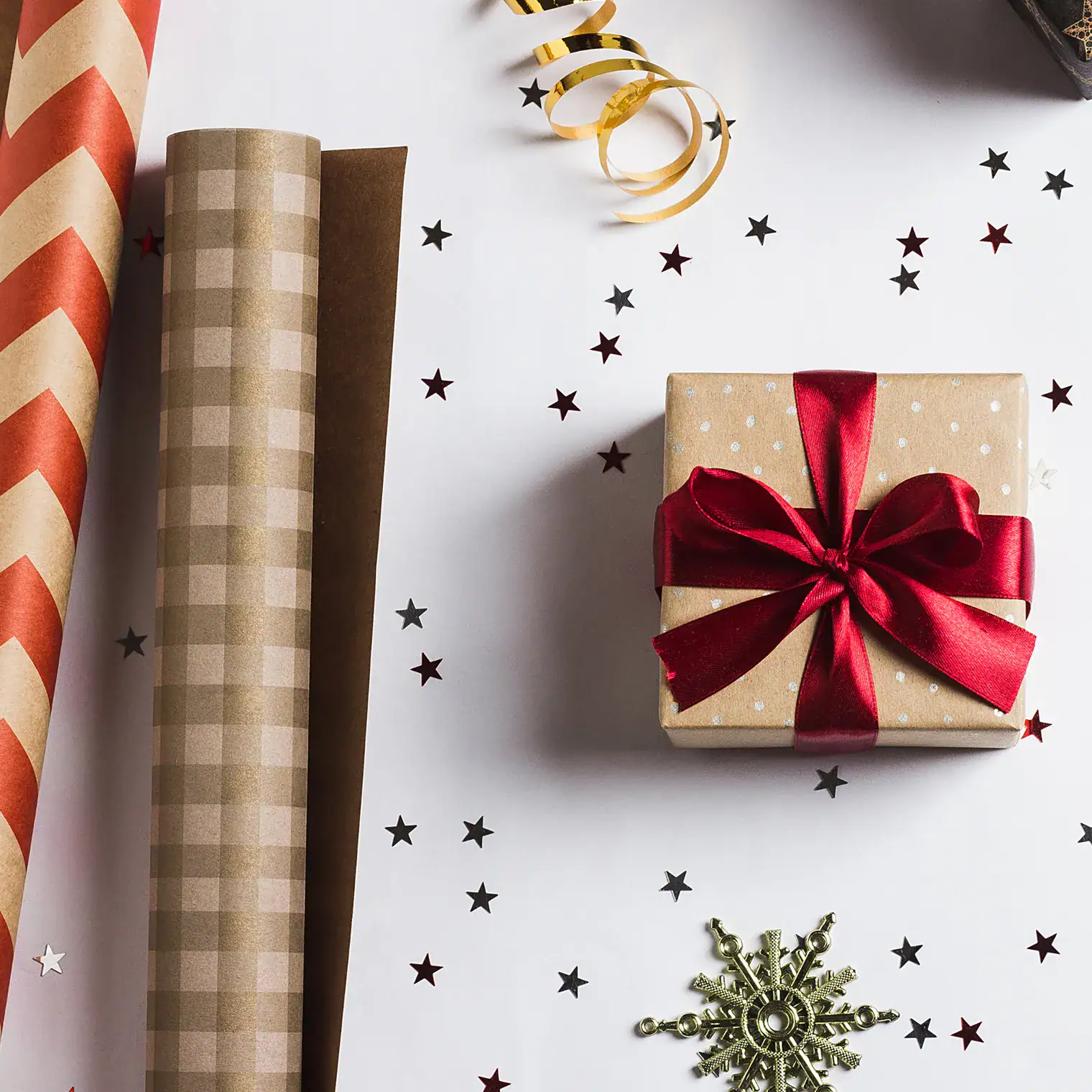 Caja de regalo envuelta en papel con lazo rojo junto a rollos de papel decorativo y adornos navideños.