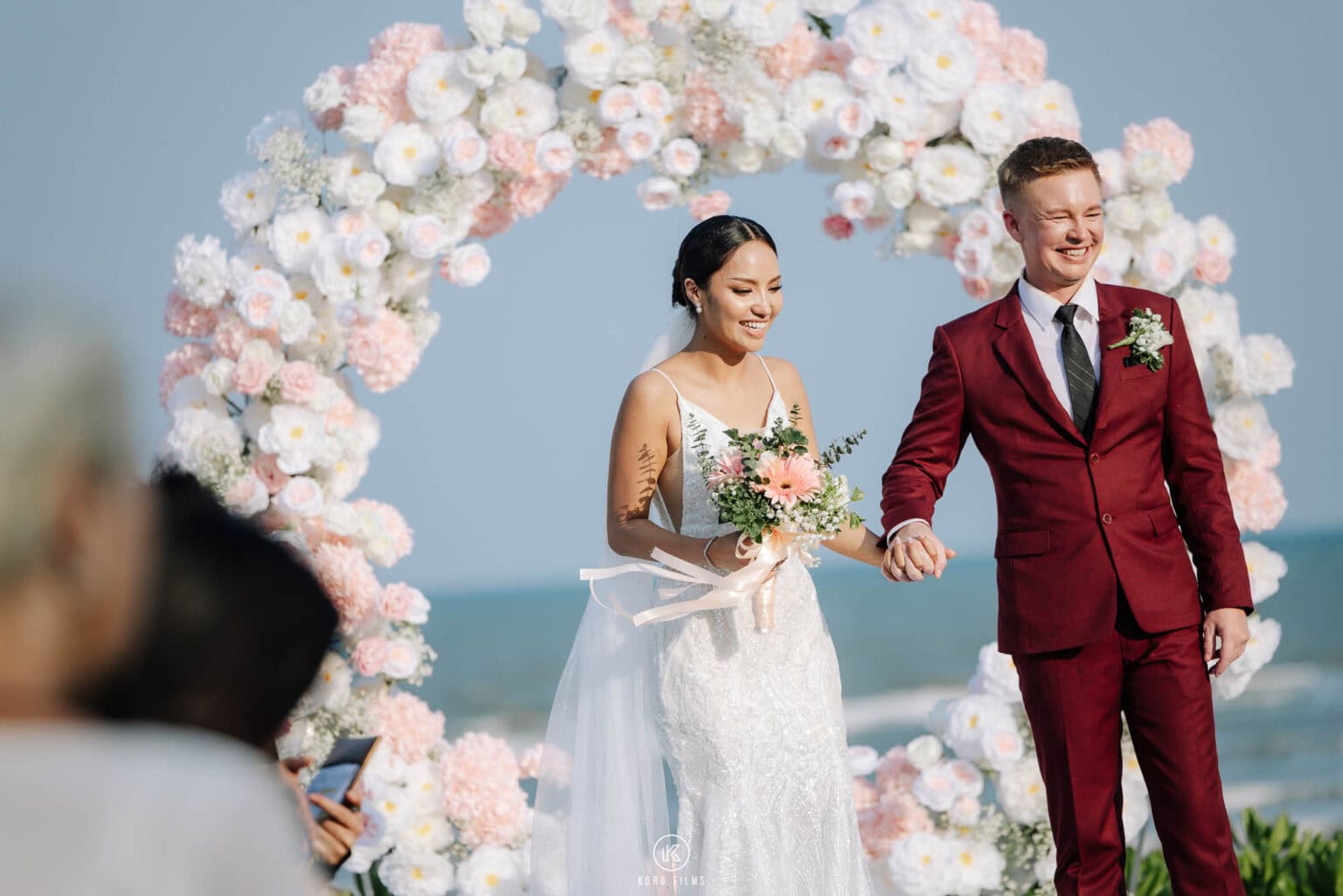 Jakub and Clear walk back down the aisle hand in hand in front of a pink and white floral ring by the sea at Sheraton Hua Hin