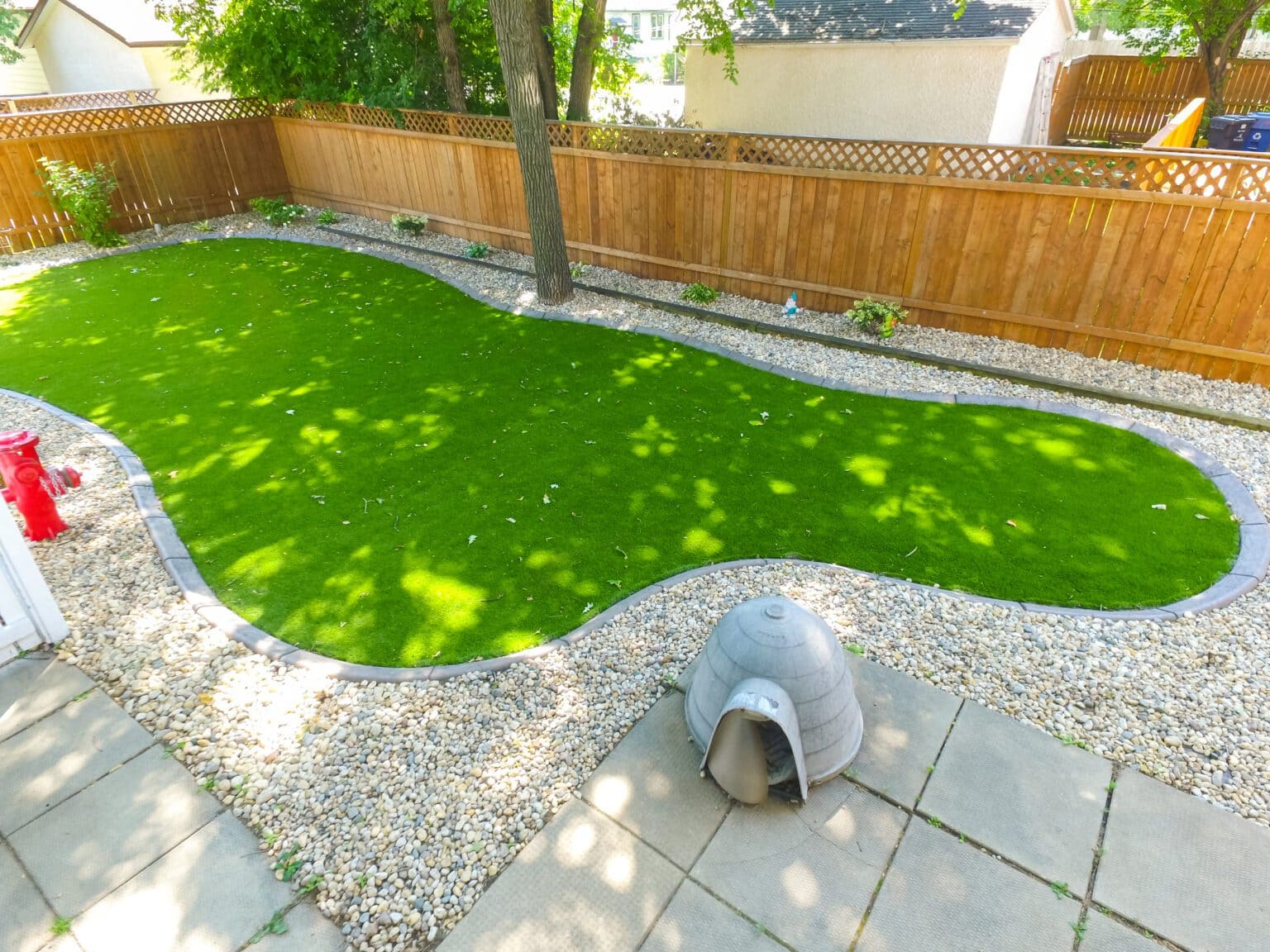 Backyard with kidney-shaped artificial grass, surrounded by decorative stones, wooden fence, and a small dog house.