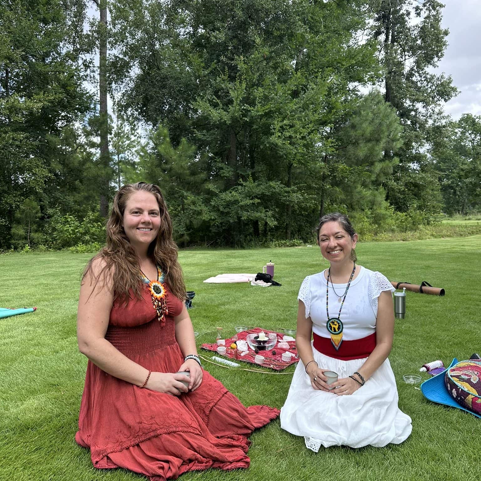 Peaceful outdoor yoga session with two women in bohemian inspired attire, practicing meditation on lush green grass surrounded by tall trees, promoting wellness, mindfulness, and holistic living.