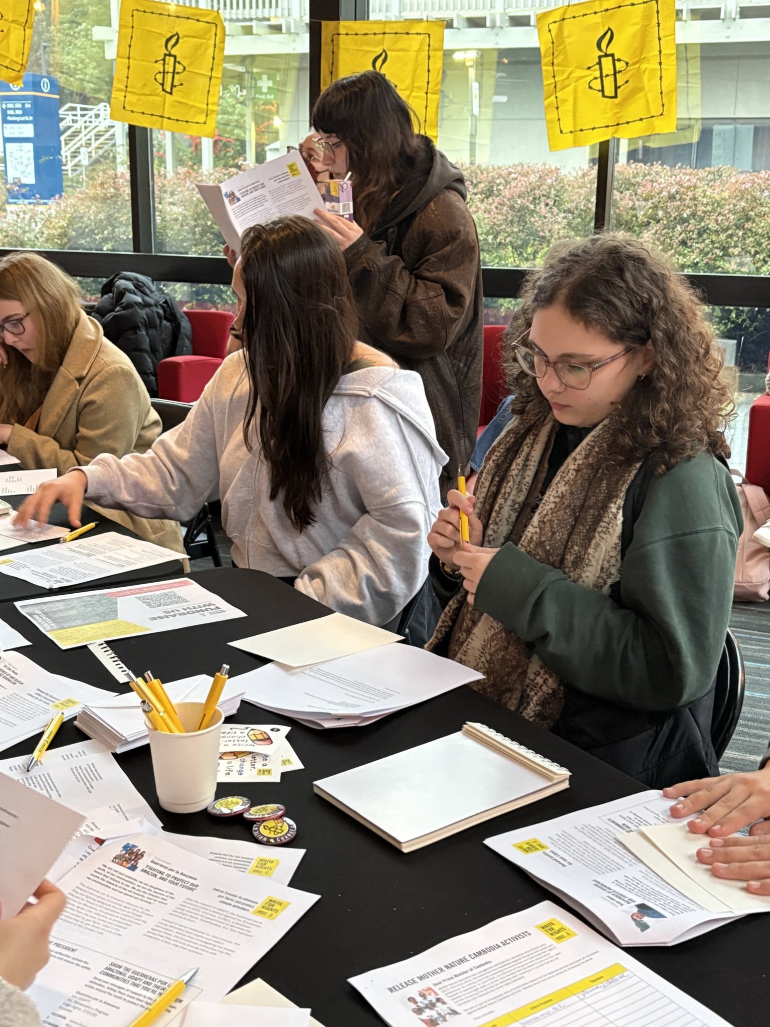 University students gathered around a table, reading case sheets and preparing letters at an Amnesty International Write for Rights event, with yellow Amnesty banners displayed behind them.