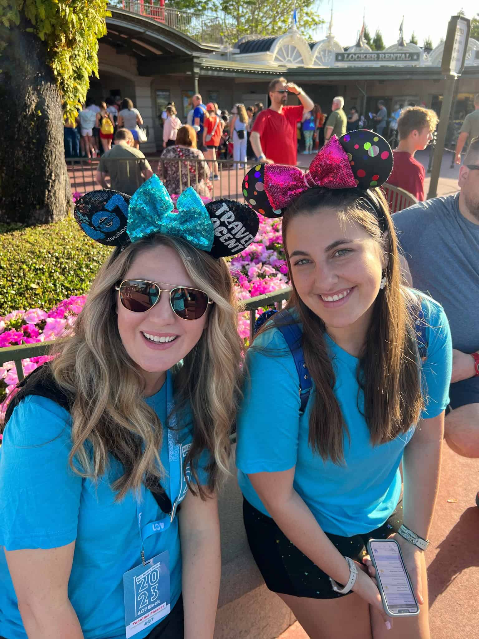 Minnie Mouse ears hats worn by two smiling women at an amusement park, with colorful floral background and crowd in the distance.