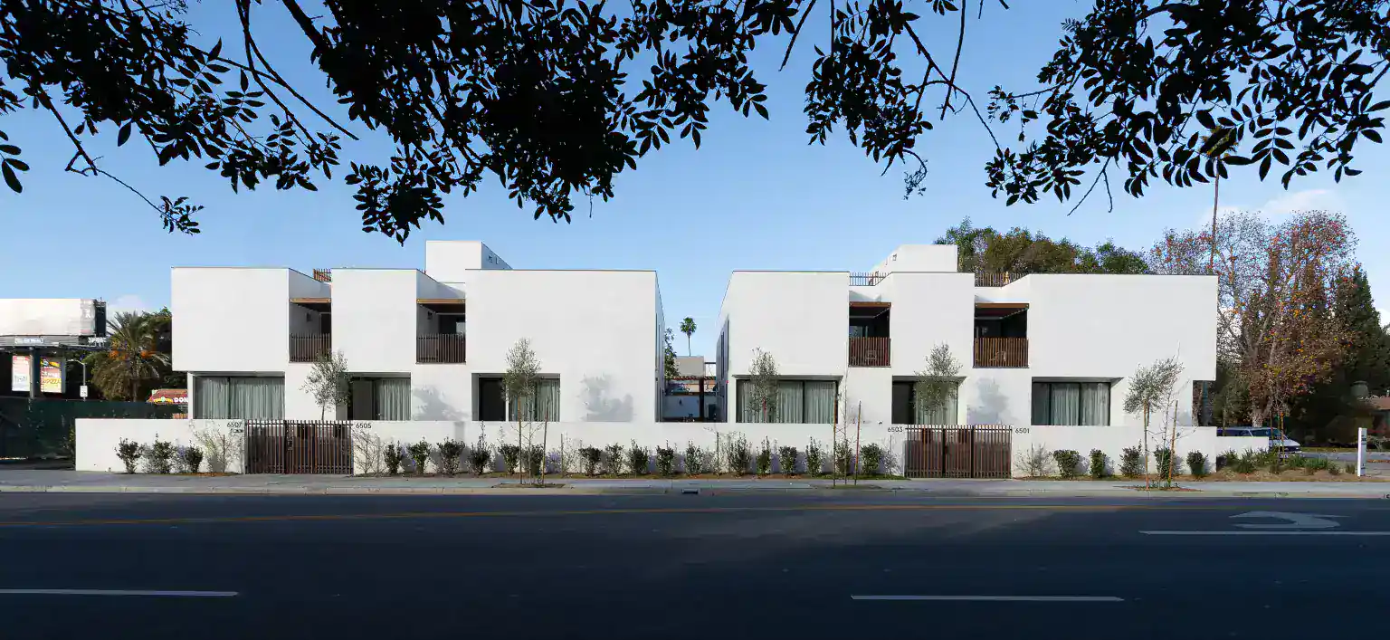 Contemporary white residential buildings with minimalist cubic forms. Wooden balcony railings and large glass doors enhance modern clarity. Sleek landscaping with small trees lines the perimeter.