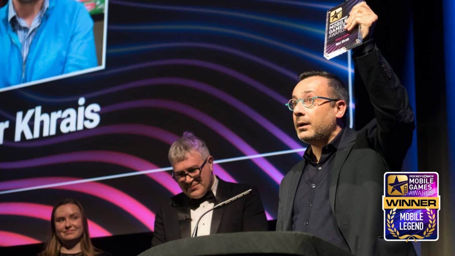 Nour Khrais, A man at a podium holds up an award, with two people standing behind him. The screen behind displays the name Khrais and a Mobile News Winner Mobile Legend badge is visible in the corner.
