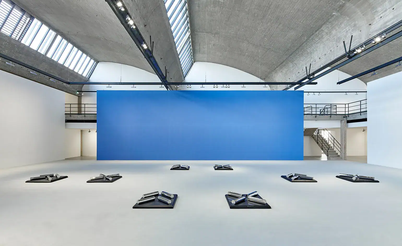 Sala espositiva principale di Gagosian Gallery Le Bourget: pavimento in resina poliuretanica Mapefloor PU 410 grigio chiaro, superficie continua senza fughe sotto soffitto a volta in cemento a vista con lucernari — installazione scultorea su basi nere con grande fondale blu, Prima Pavimenti