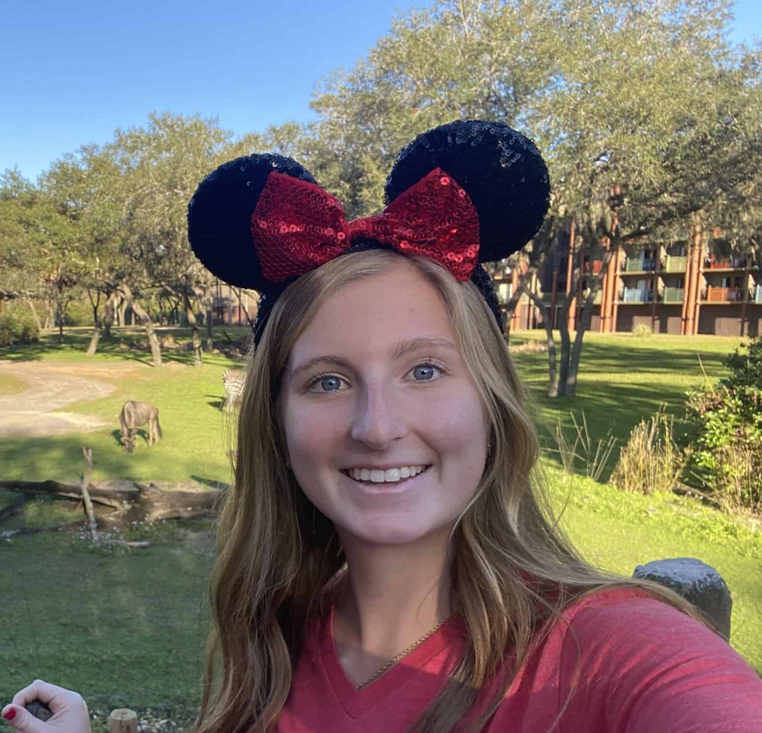 Young woman wearing Minnie Mouse ears taking selfie at zoo with animals in background.