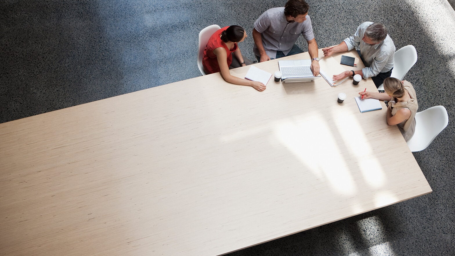 Four people sit around a large wooden table, engaged in discussion. A laptop, notepad, and coffee cups are on the table.