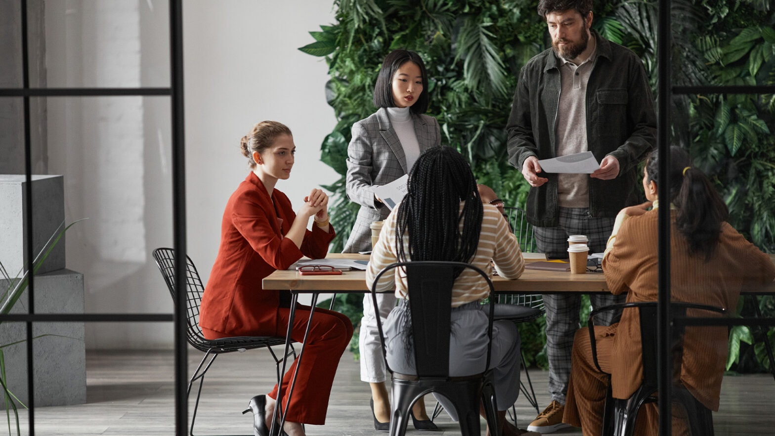 Six people in a conference room with a plant wall, engaged in a meeting. Some are seated, while others stand holding papers.