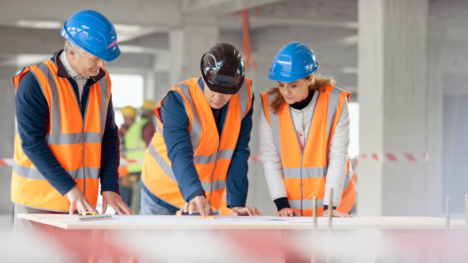 Three construction workers in orange vests and hard hats examine a blueprint on a table at a building site.