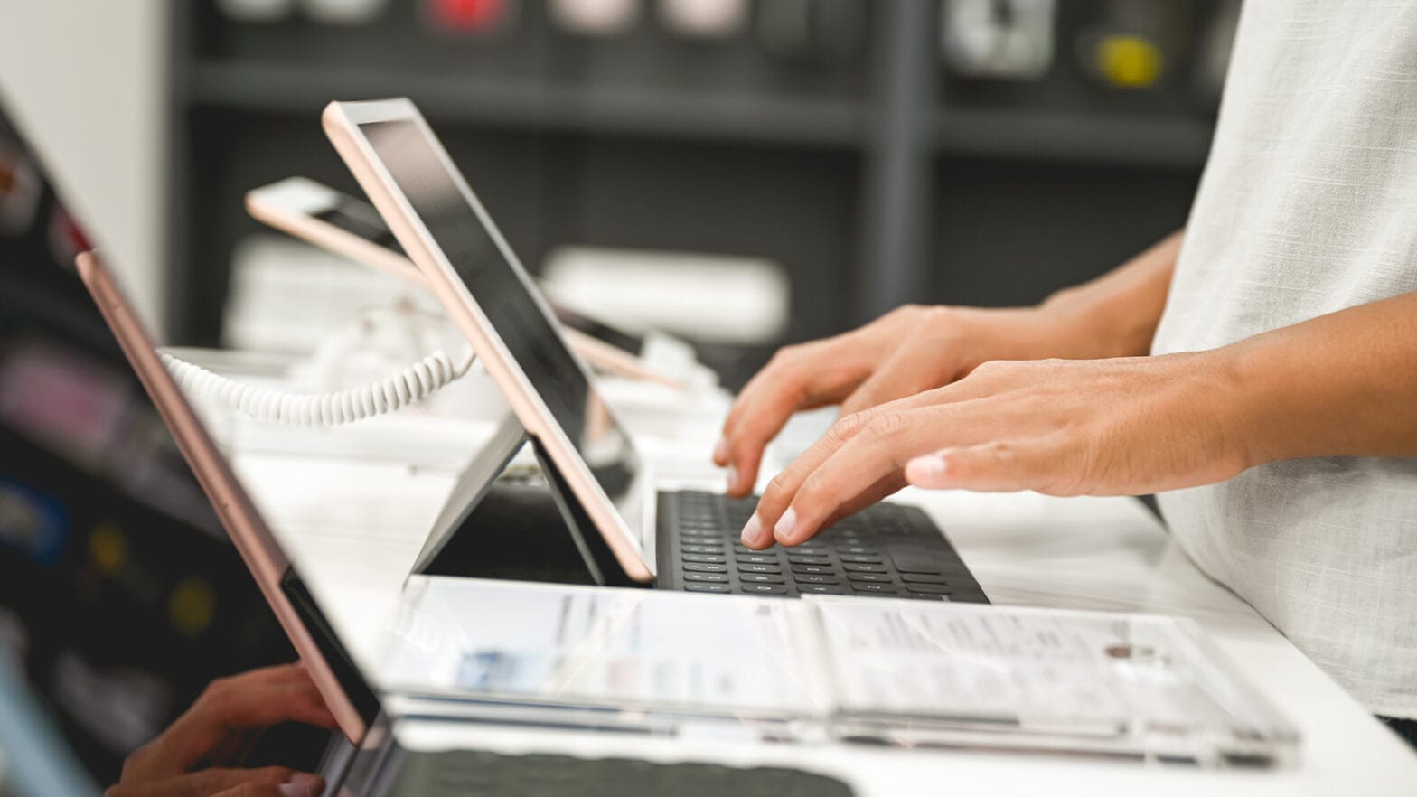 Person typing on a keyboard connected to a tablet in a tech store, with various devices displayed on the counter.