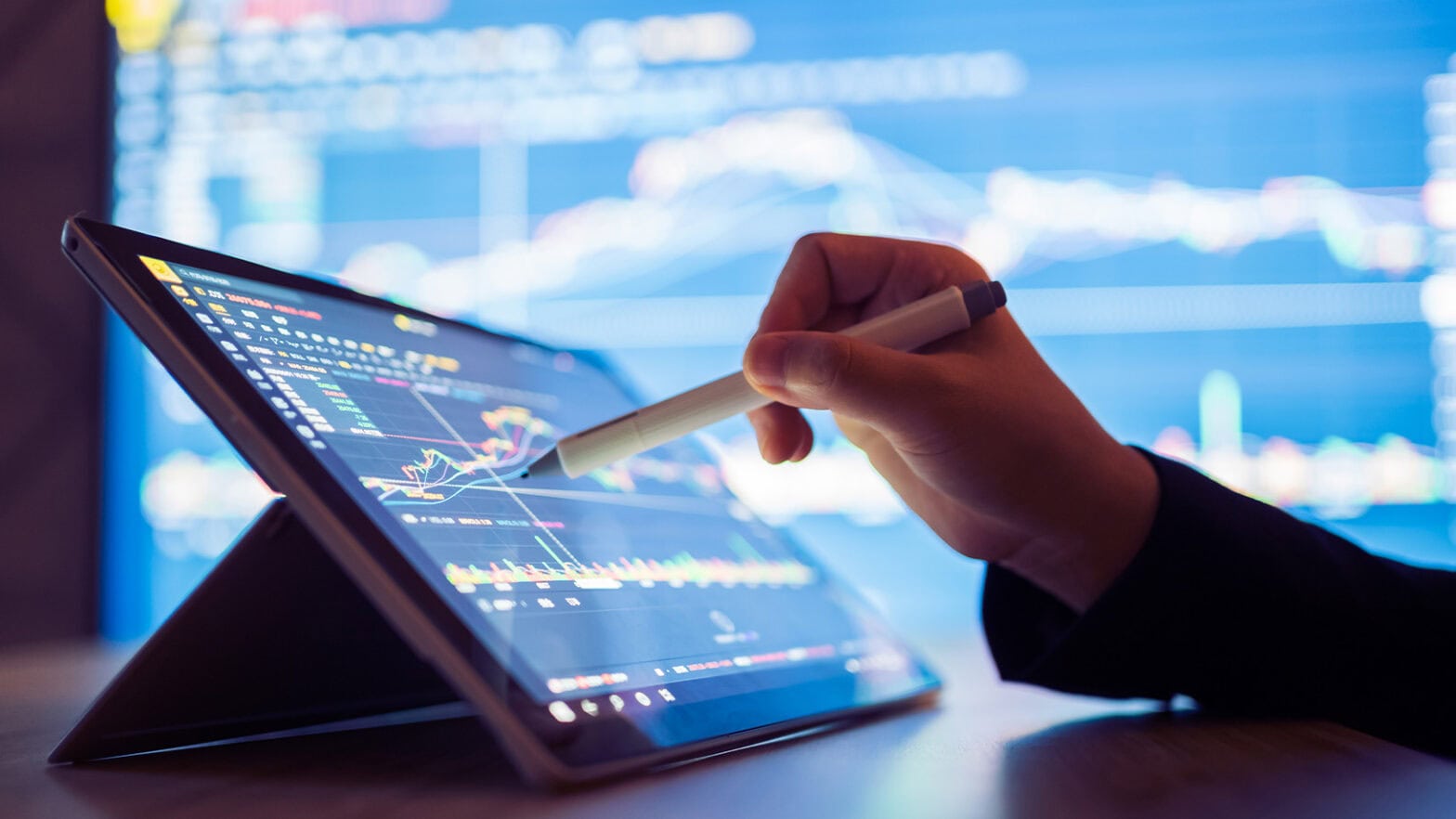 Person using a stylus to interact with a tablet displaying financial graphs, with a large screen showing more charts in the background.