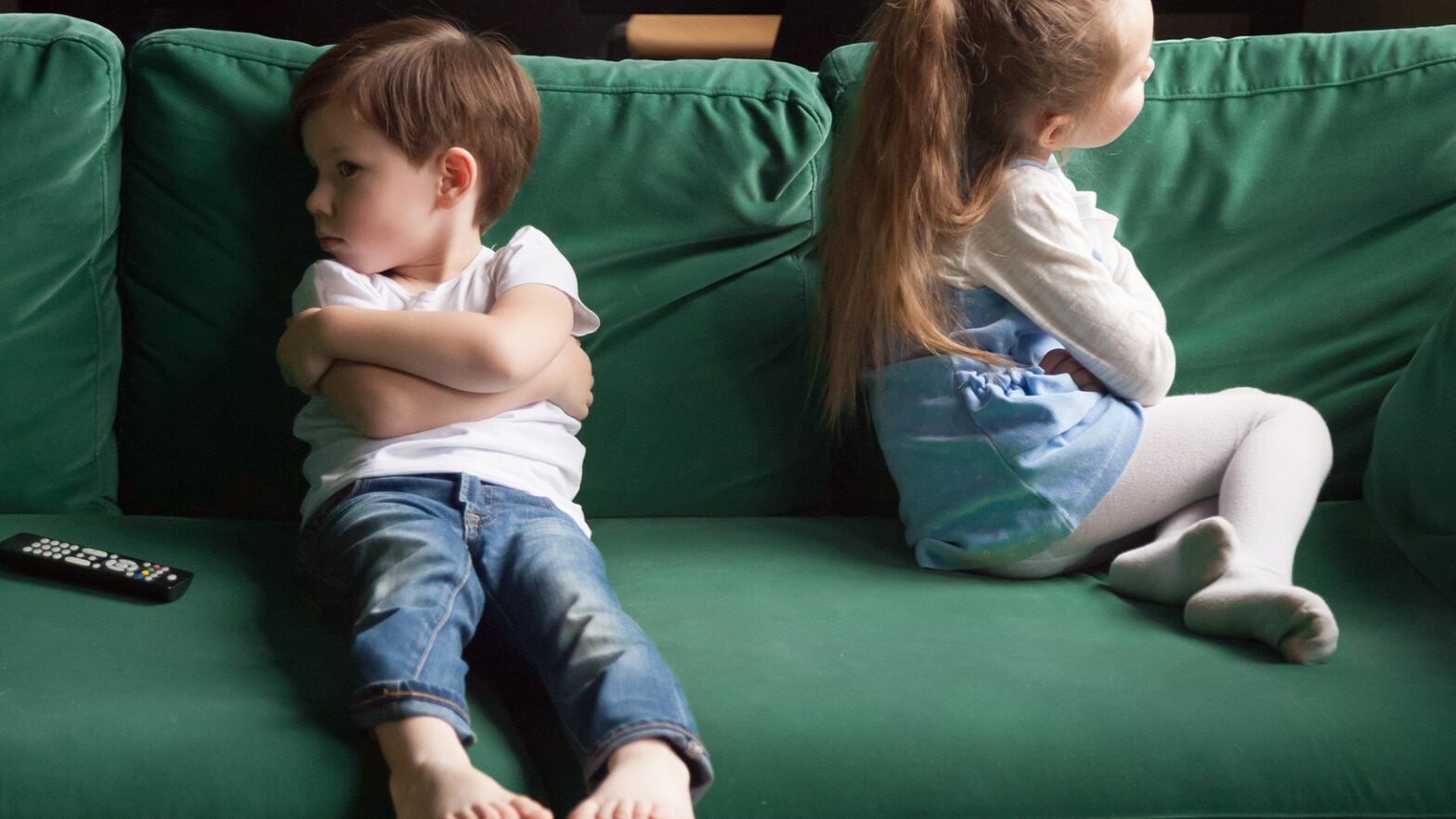 A boy and a girl sit back-to-back on a green couch with arms crossed, facing away from each other. A remote control is on the couch.