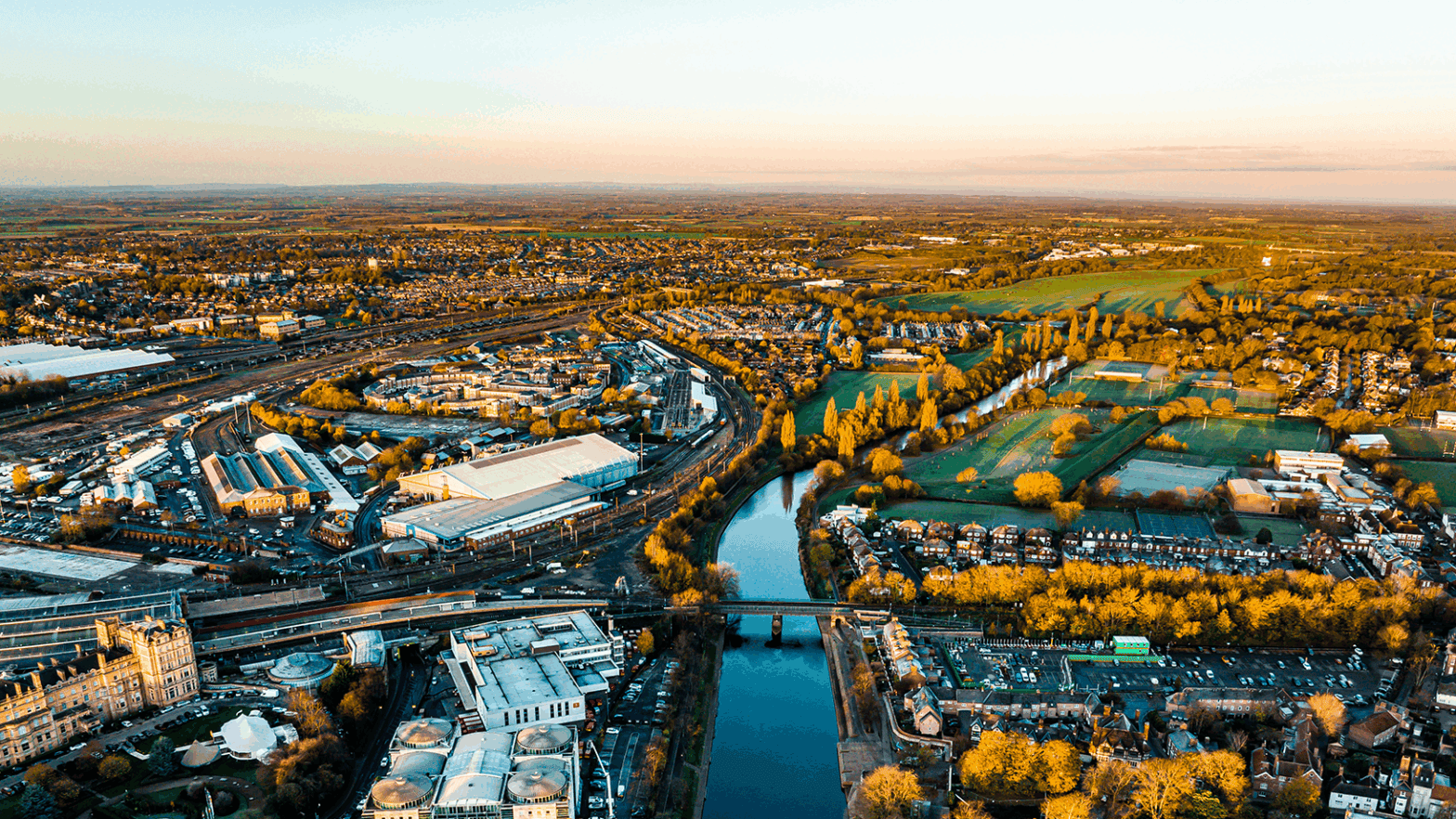 Aerial view of a river running through a city with buildings, roads, and green fields at sunrise or sunset.