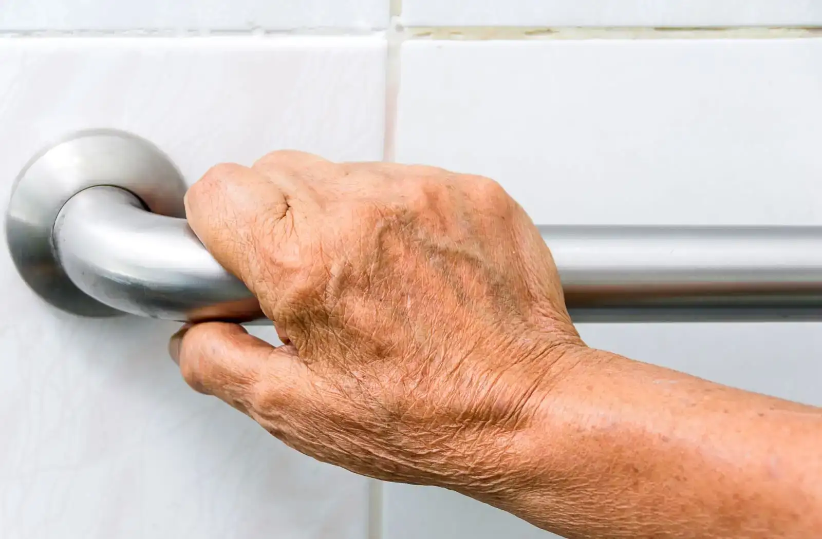 Close-up of an elderly person's hand gripping a stainless steel grab bar in a bathroom, demonstrating accessibility and support for individuals with mobility challenges.