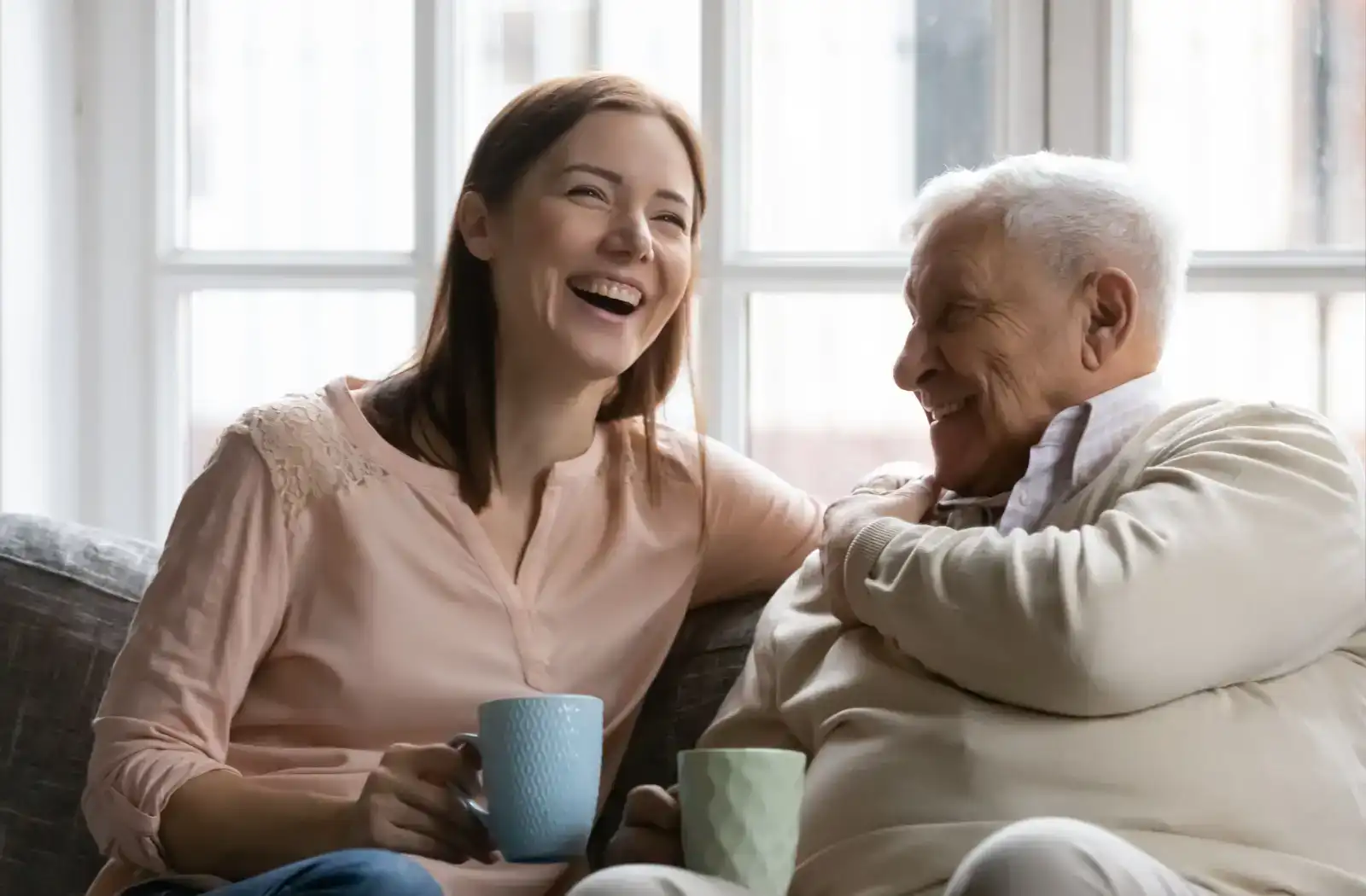 A woman and an elderly man share a joyful moment while drinking tea in a cozy living room, highlighting the warmth of intergenerational relationships.