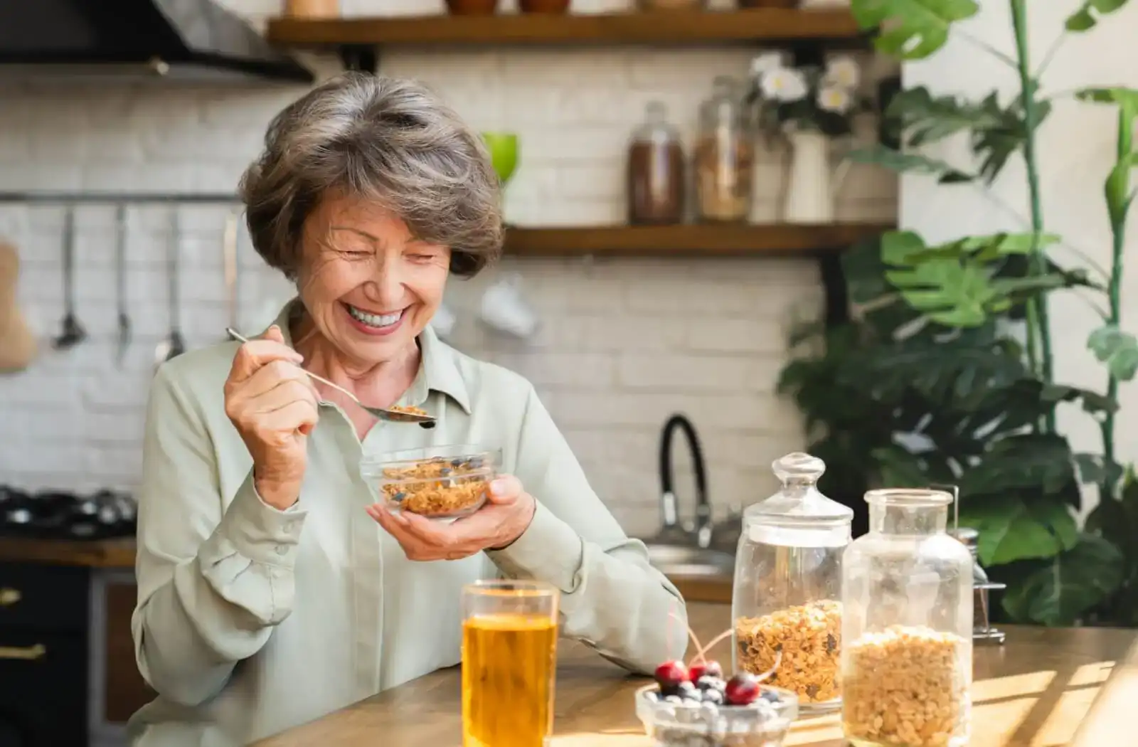 Smiling elderly woman enjoying a bowl of granola at a cozy kitchen table, with a glass of iced tea and jars of ingredients in the background.