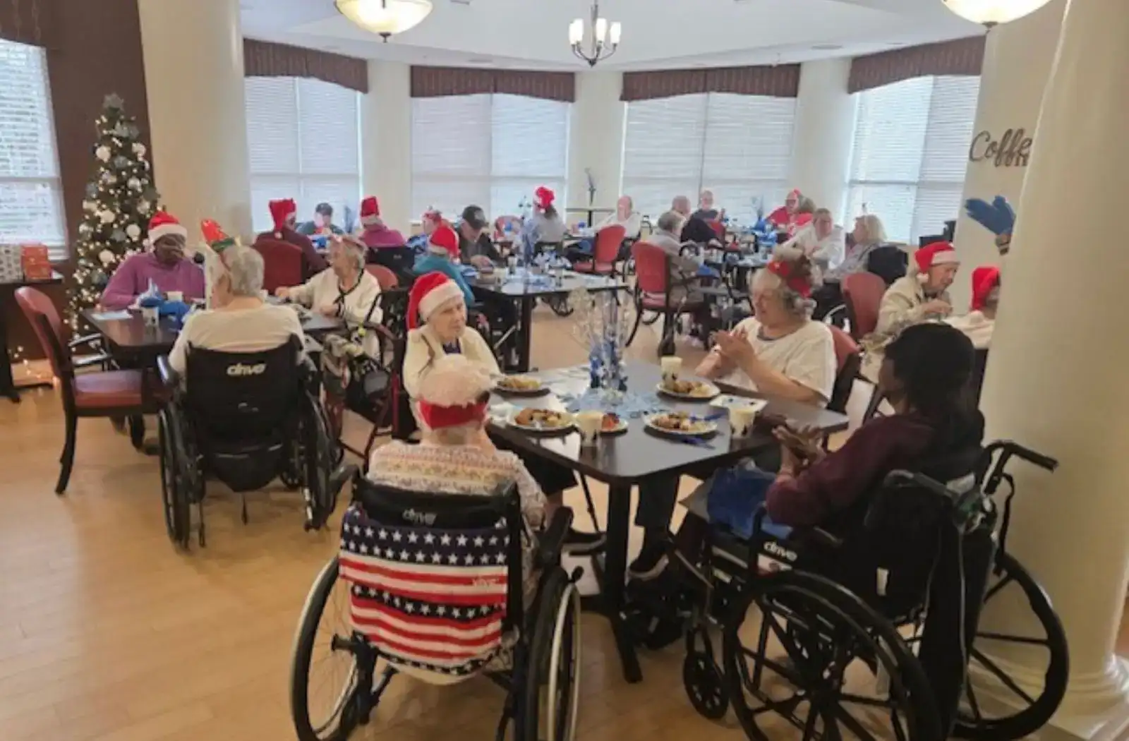 Residents enjoying a festive meal in a dining hall, wearing holiday hats and seated in wheelchairs, with tables set for a communal dining experience.