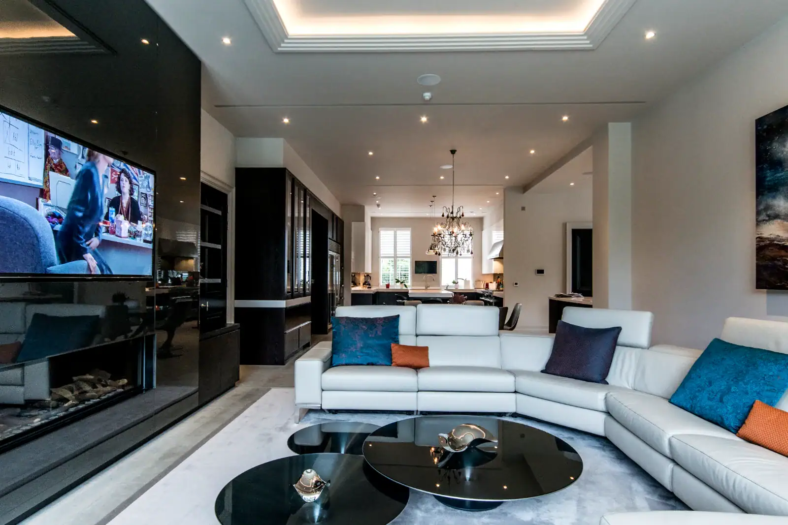 Modern living room with a white corner sofa, blue and orange accent cushions, round black coffee tables, wall-mounted TV—ideal for home cinema installation in Oxshott, Surrey—plus chandelier in the dining area and recessed ceiling lighting.