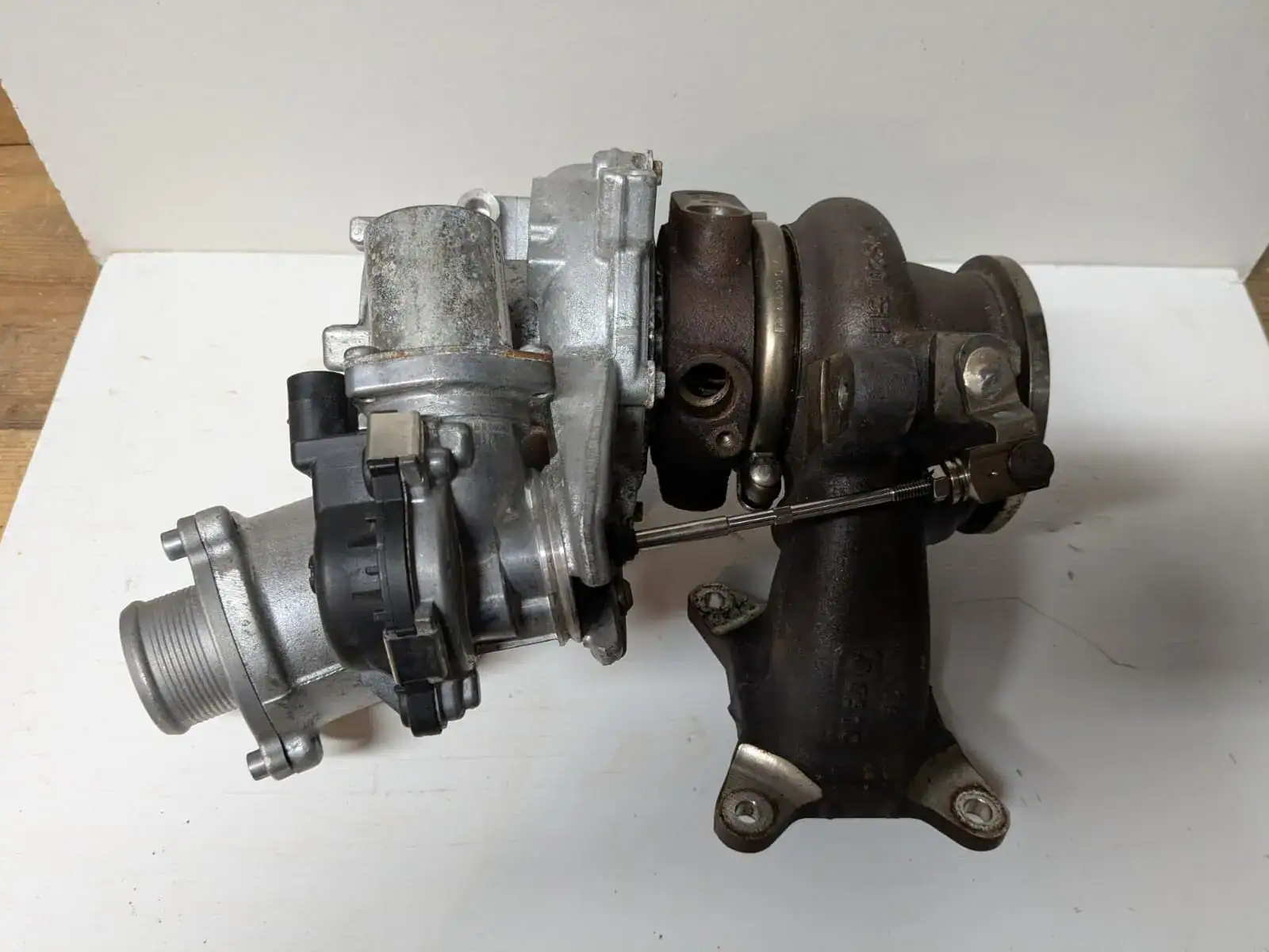 Side view of an IS20 turbocharger showing aluminum compressor housing, electronic wastegate actuator, linkage rod, and cast turbine housing