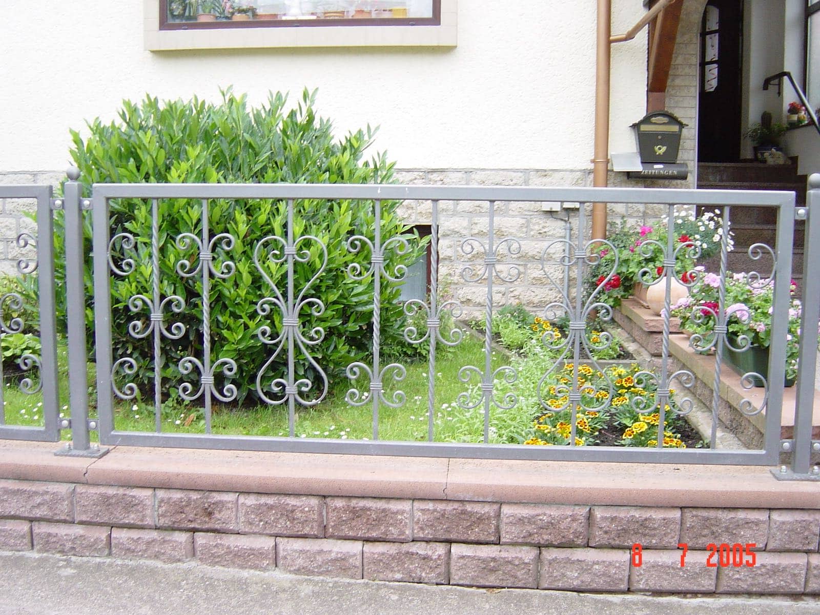 Grauer dekorativer Metallzaun mit verschnörkelten Ornamenten und gedrehten Stäben auf einer Ziegelmauer, im Garten vor einem Haus. Qualität von Zaunbau von Stahl- und Metallbau Drost.