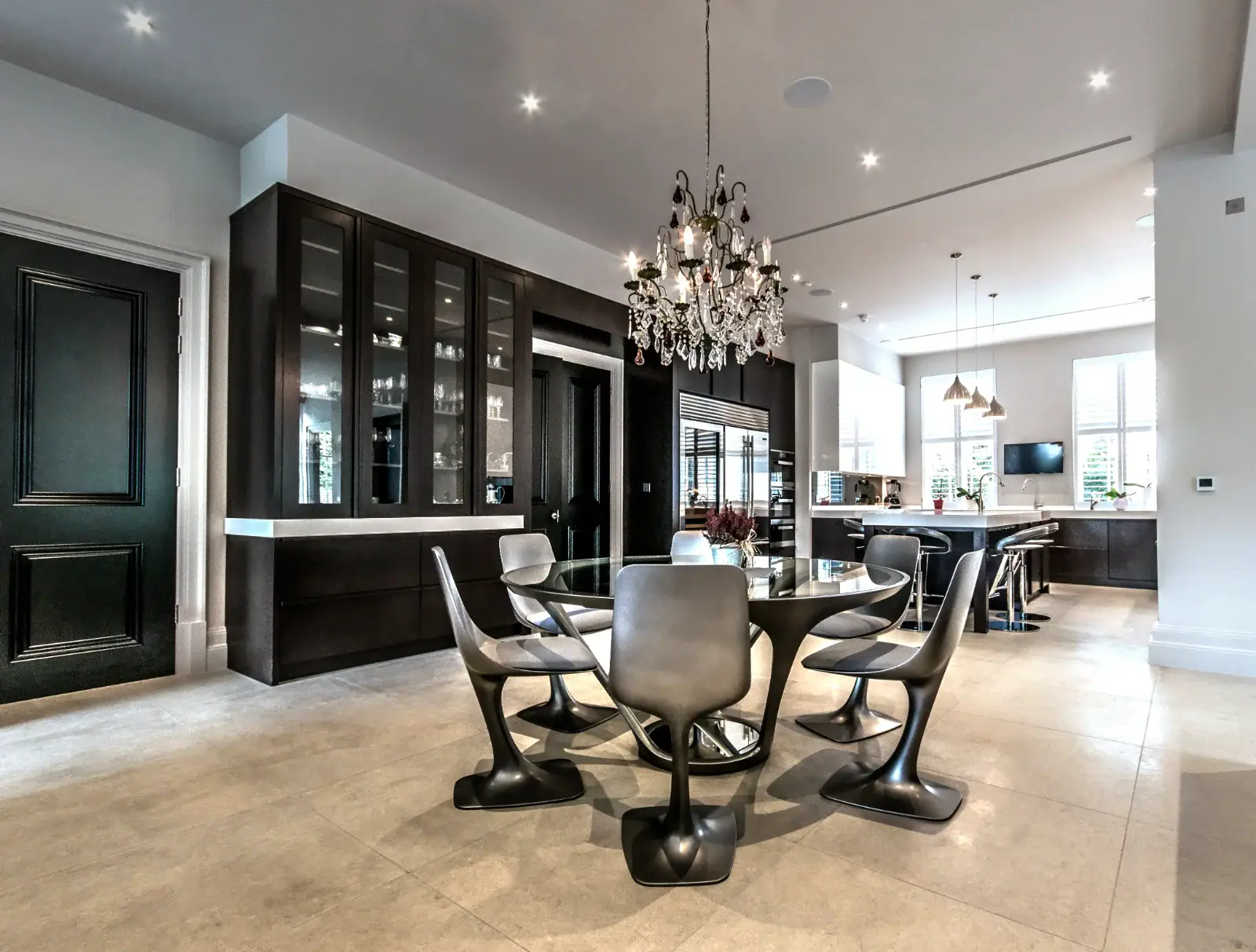 Modern open-plan kitchen and dining area with sleek black cupboards, a glass dining table with six grey chairs, chandelier above the table, large windows letting in natural light, and smart lighting installation in Oxshott Surrey.