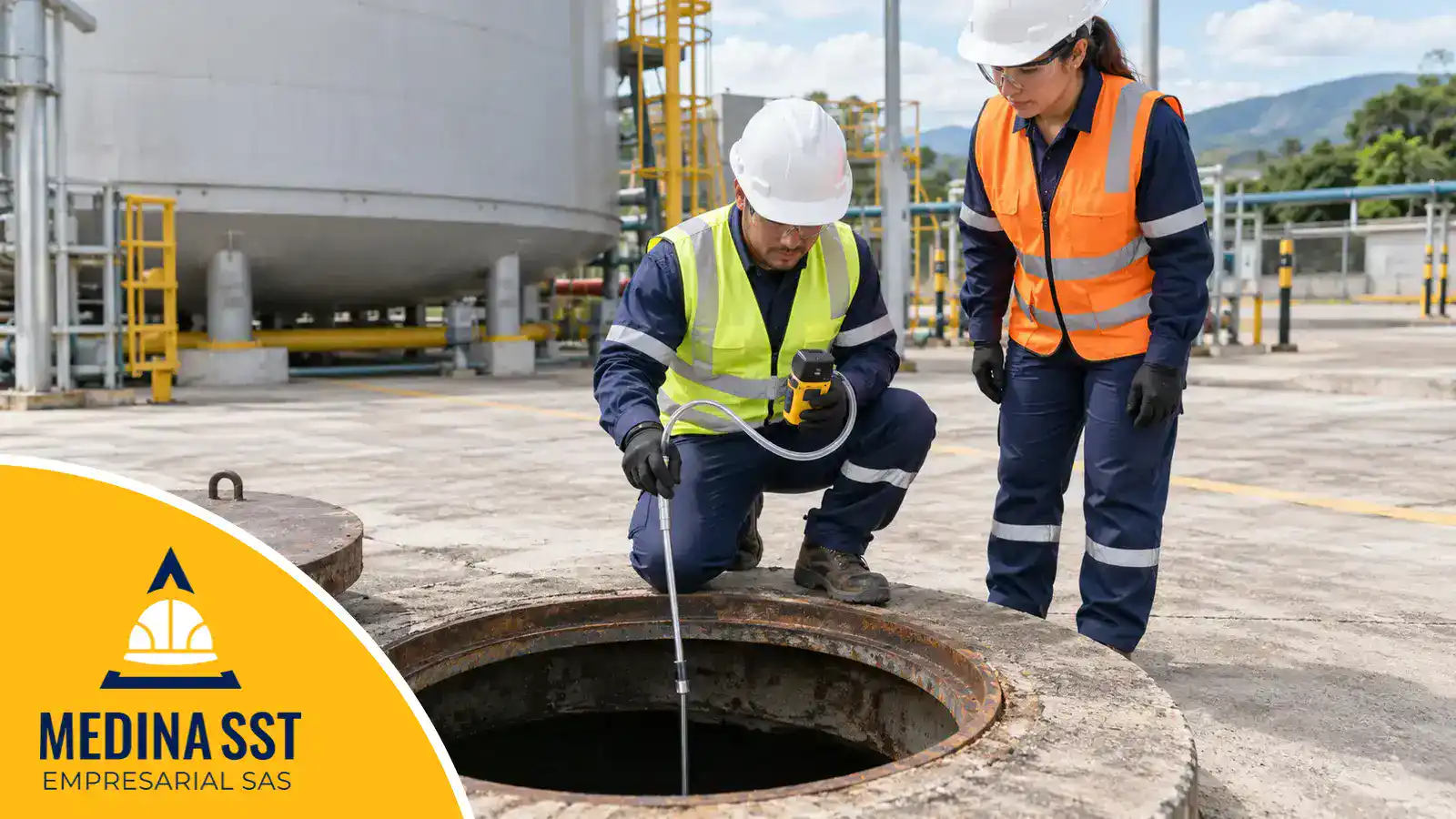 Técnico con casco, gafas y chaleco reflectivo, arrodillado junto a la boca abierta de un tanque, mide la atmósfera del fondo con un detector de gas portátil, su manguera de muestreo y la sonda metálica