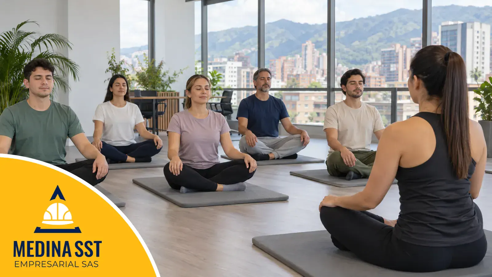 Instructora de yoga empresarial sentada frente a trabajadores que siguen la sesión sentados en colchonetas, en una sala de oficina con ventanales