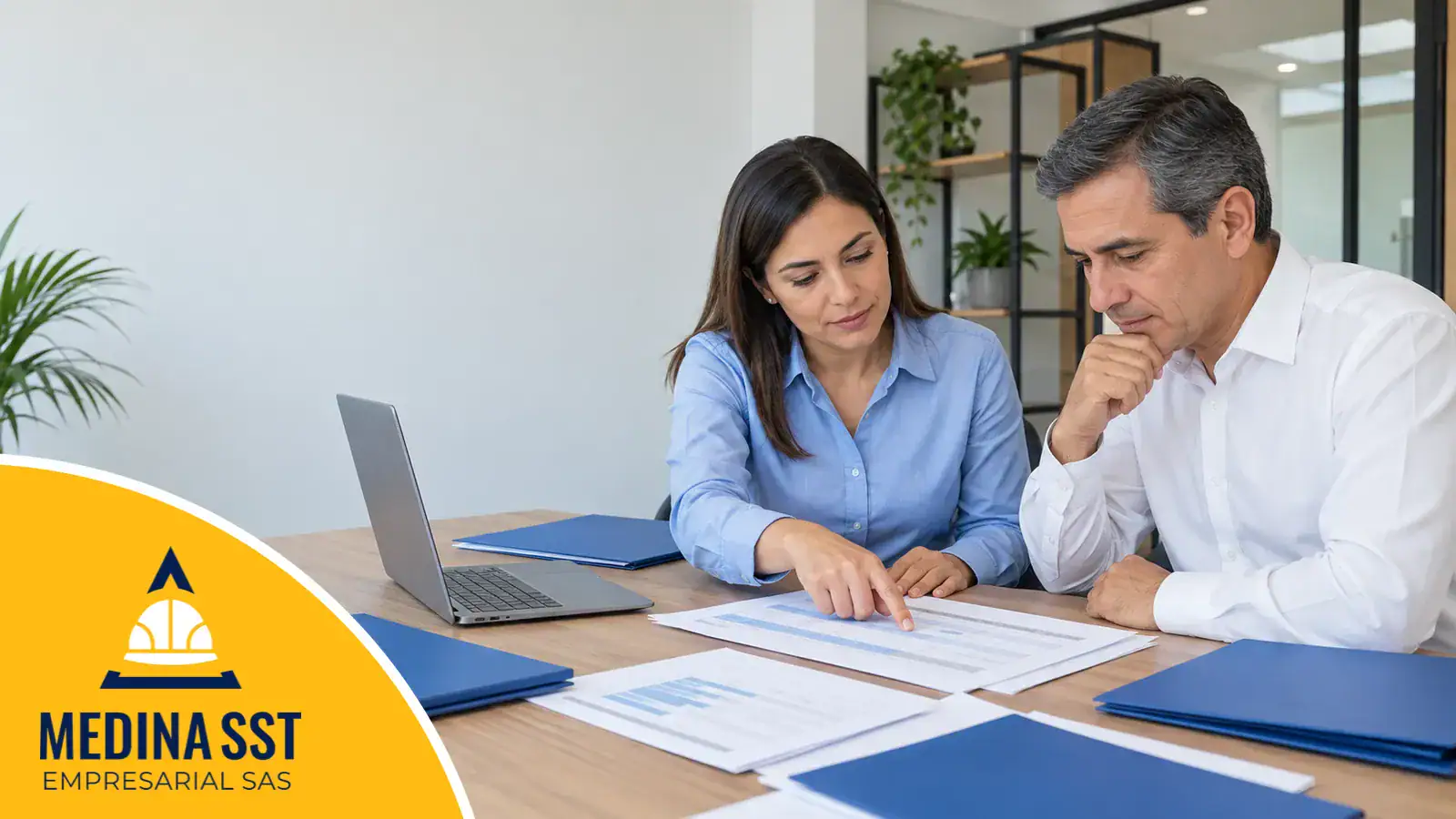 Matriz de cumplimiento legal: una profesional de seguridad y salud en el trabajo señala una tabla impresa al gerente de la empresa sobre un escritorio con un portátil y carpetas azules