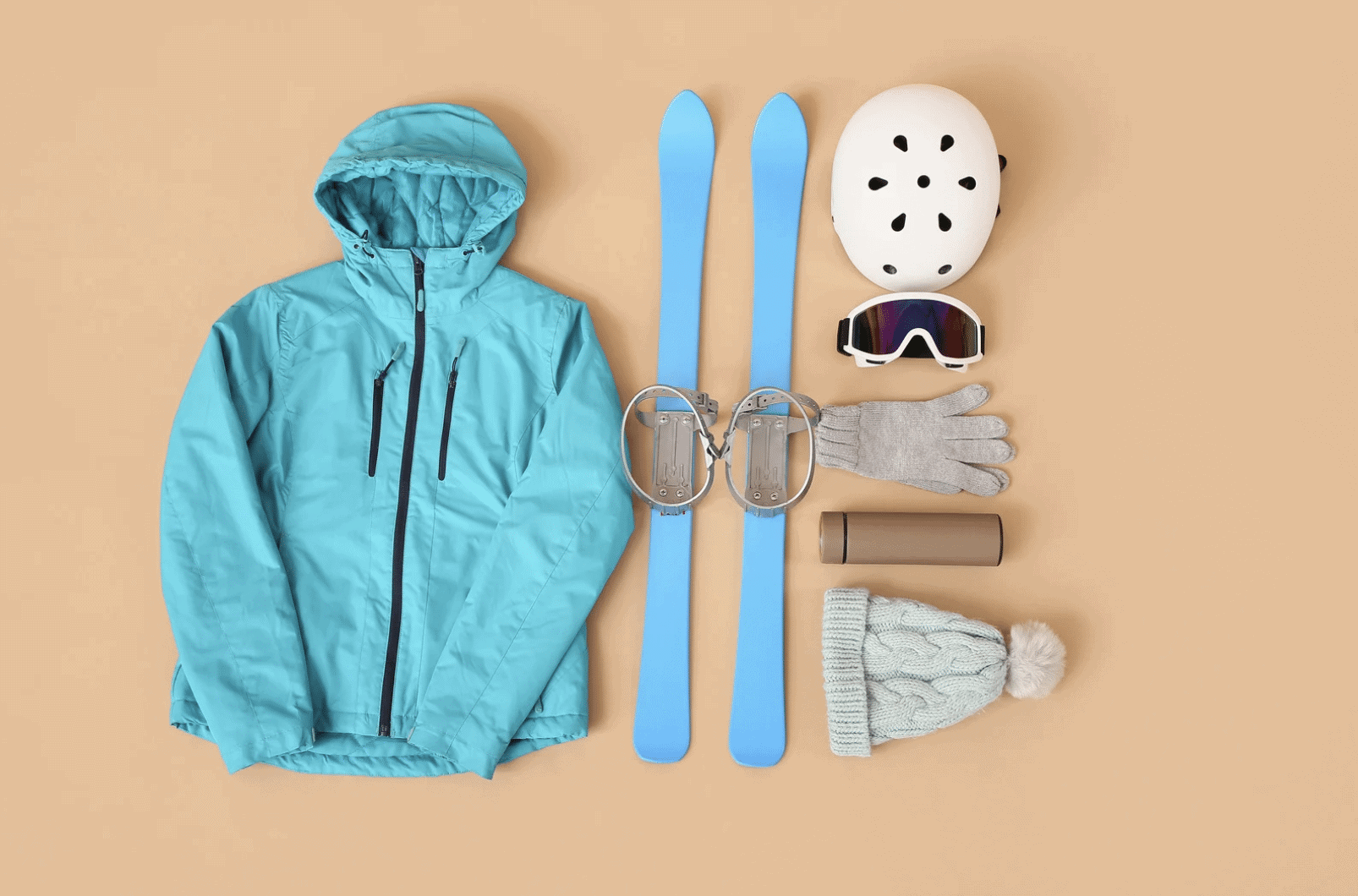 Flat lay of ski trip packing list essentials including a teal ski jacket, blue skis with bindings, white helmet, ski goggles, gloves, knit hat, and thermos on a neutral background.