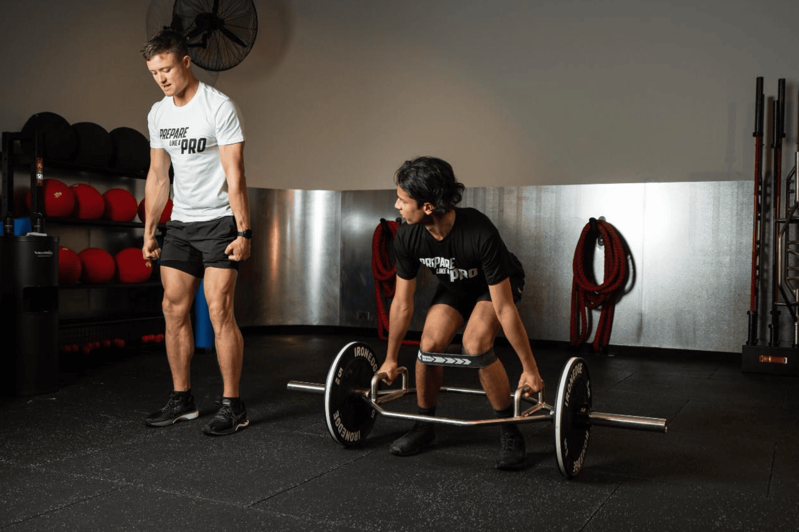 One man lifting a barbell and another watching him in a well-equipped gym, both wearing 'PREPARE LIKE A PRO' shirts.