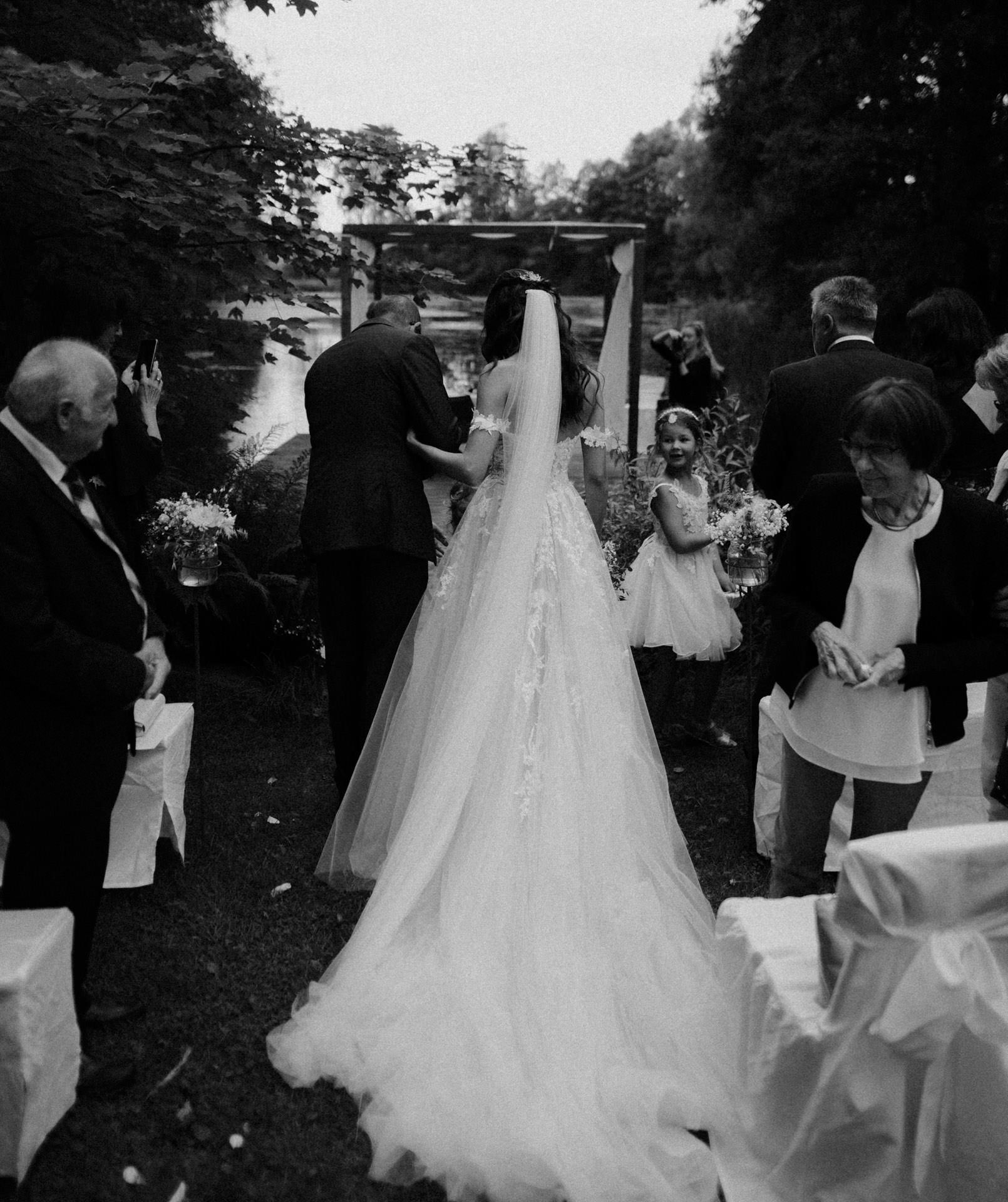 Das Schwarzweißfoto eines Hochzeitsfotografen aus Regensburg zeigt Braut und Bräutigam, die im Freien umgeben von Gästen einen Gang entlanggehen. Die lange Schleppe und der Schleier der Braut ziehen hinterher, während ein junges Blumenmädchen ihnen folgt. Bäume und Stühle bilden die bezaubernde Kulisse für diesen wunderschönen Moment.