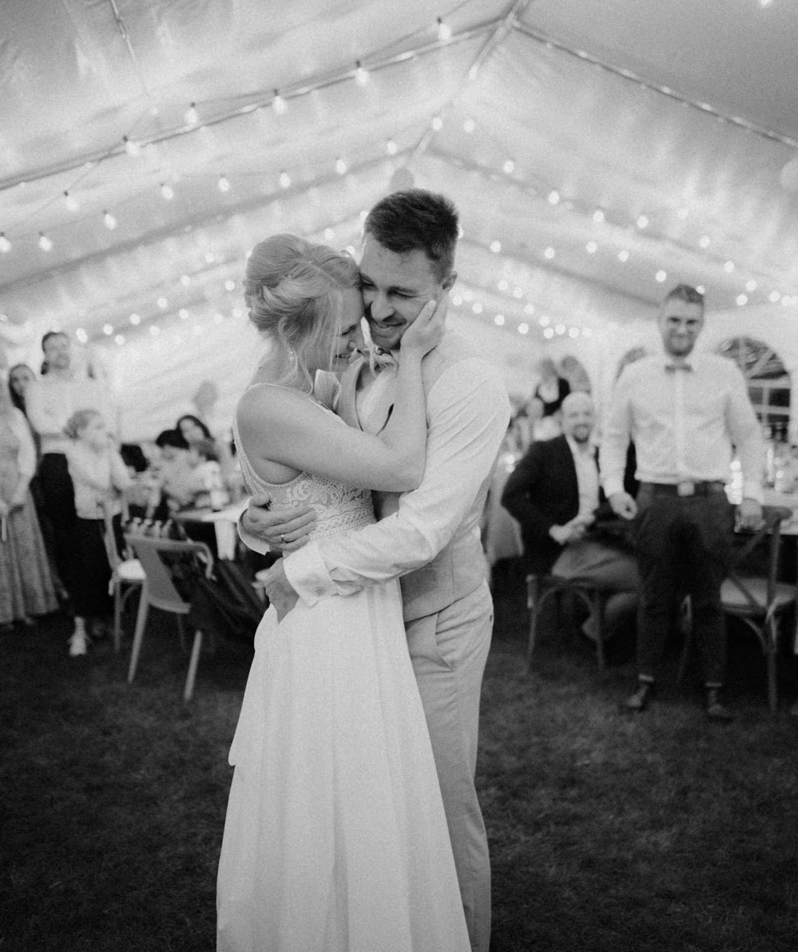 Ein Paar teilt einen zärtlichen Moment während seines ersten Tanzes auf einer Hochzeitsfeier in einem mit Lichterketten geschmückten Zelt. Im Hintergrund sitzen die Gäste an Tischen und beobachten das Paar lächelnd, während der Hochzeitsfotograf Regensburg jede kostbare Sekunde festhält.