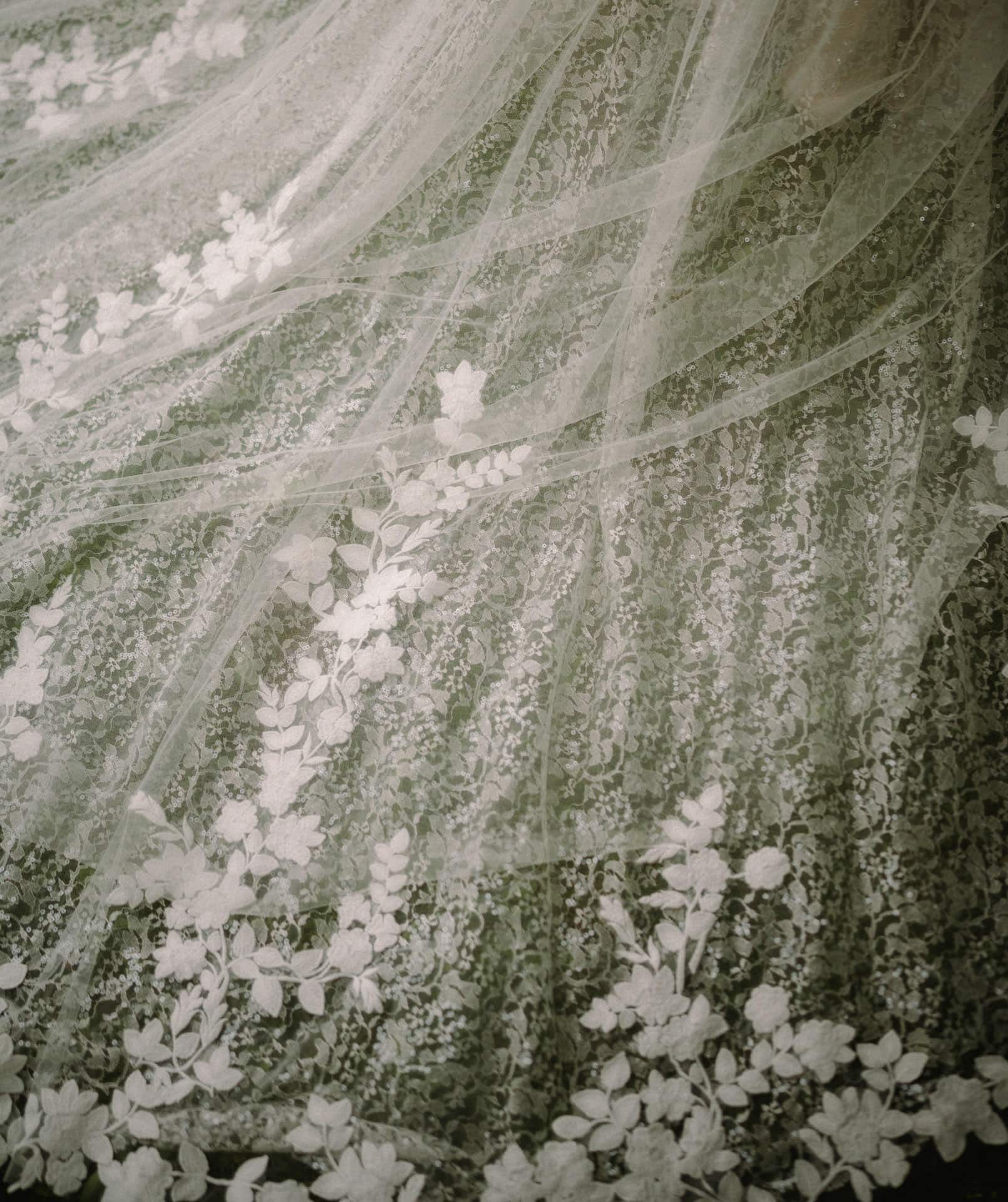 Nahaufnahme eines zarten, weißen Spitzenstoffs mit Blumenmotiven und aufwendigen Blatt- und Blumenmustern, perfekt für einen Hochzeitsfotografen aus Regensburg. Die Spitze ist auf einem dunkleren Hintergrund geschichtet, wodurch ein weicher, eleganter Look entsteht, der sich ideal für Hochzeiten eignet.