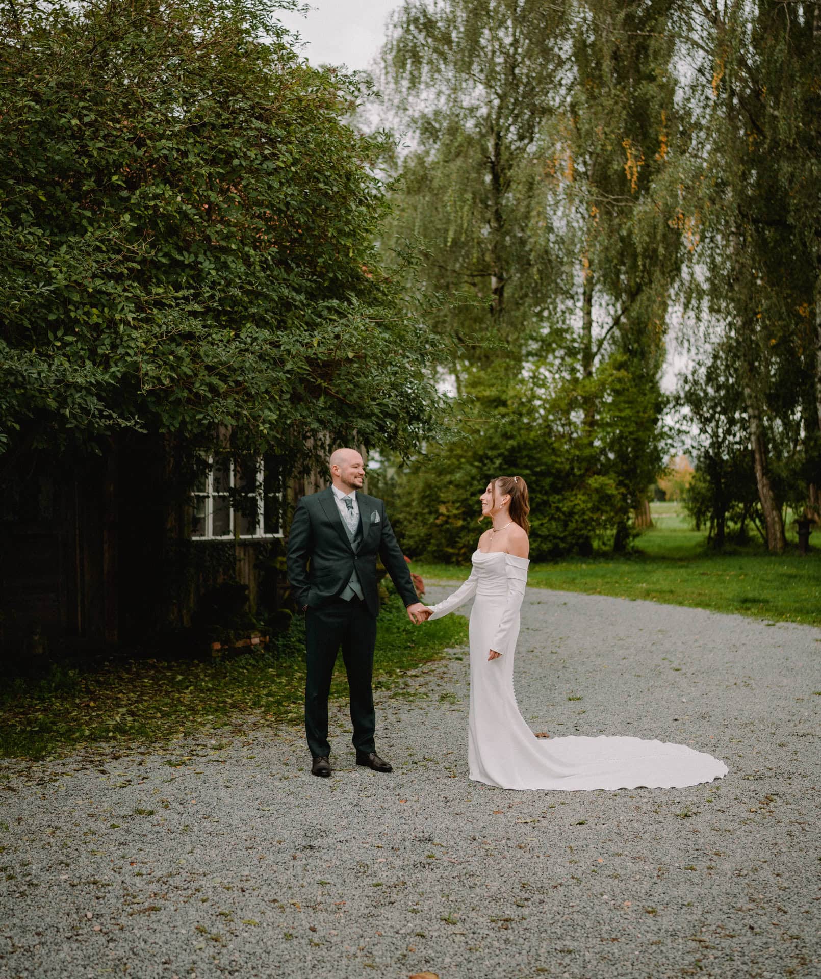 Braut und Bräutigam stehen auf einem Kiesweg, halten Händchen und lächeln sich an. Die Braut trägt ein langes, weißes schulterfreies Kleid. Aufgenommen von einem Hochzeitsfotografen aus Regensburg sind sie von üppigem Grün umgeben, im Hintergrund sind hohe Bäume zu sehen.
