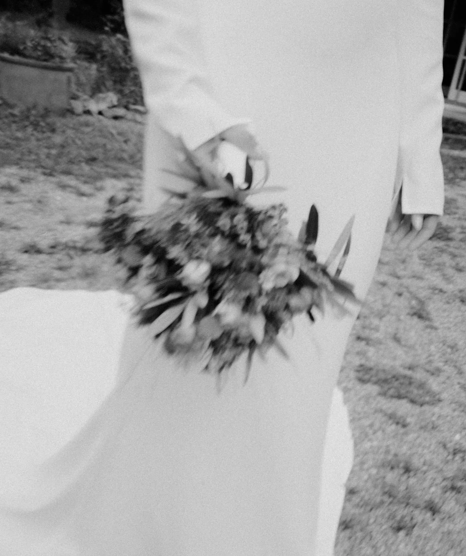 Ein Schwarzweißbild einer Person in einem langen, eleganten Kleid, die einen Blumenstrauß hält, fängt die Essenz der Romantik ein. Aufgenommen von einem Hochzeitsfotografen aus Regensburg, erzeugt die Außenkulisse mit Gras und Unschärfeeffekt eine weiche, verträumte Qualität.